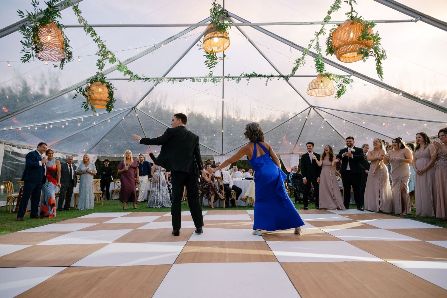 Couple sharing energetic first dance on checkered floor under elegant tent with hanging greenery.