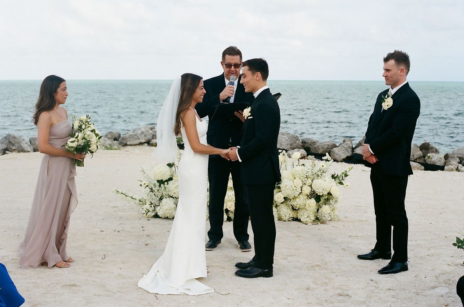 Two grooms exchange vows at a beachside wedding ceremony with ocean views, officiant, best man, and bridesmaid present.