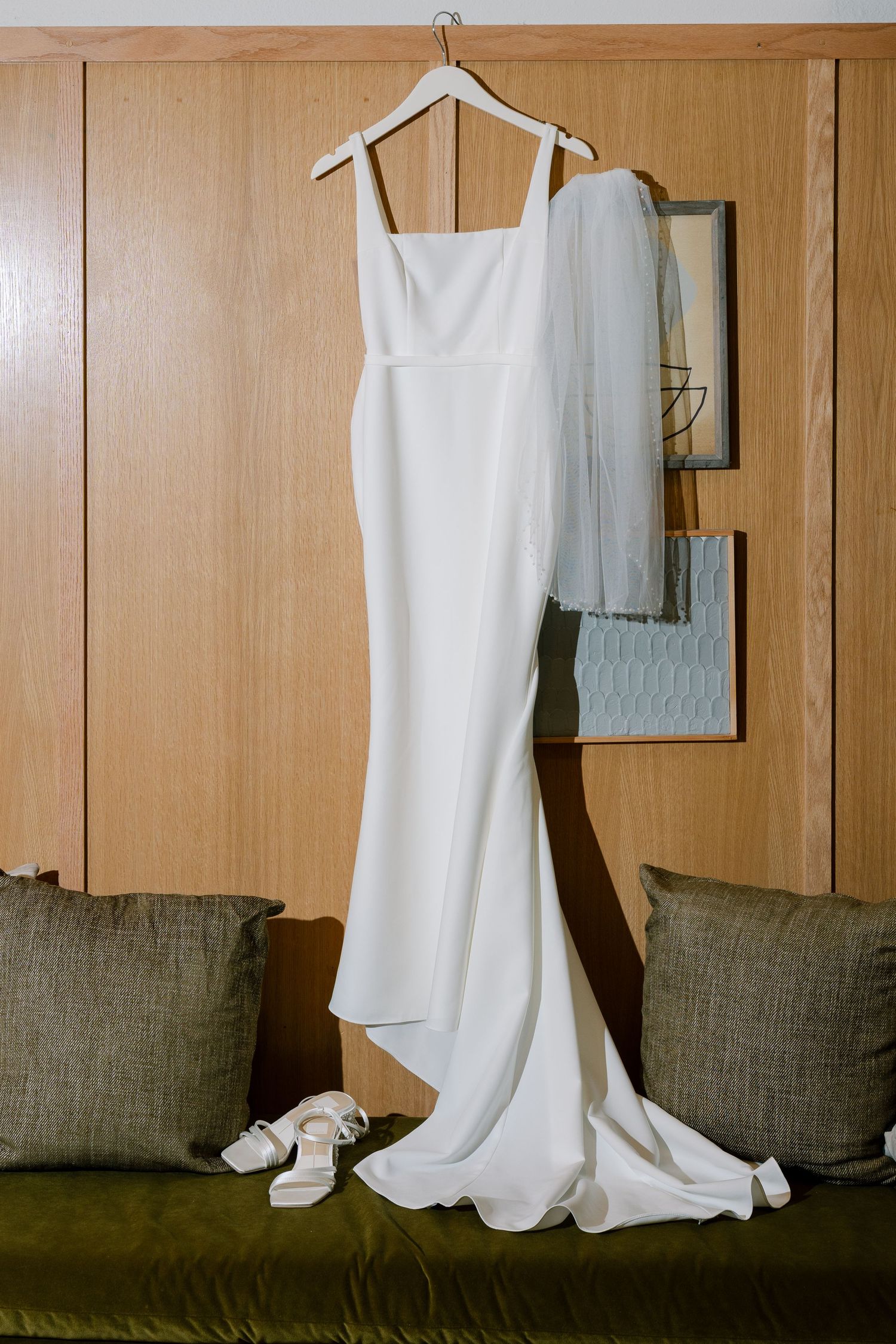 Elegant white wedding dress with square neckline hanging on wooden wall with veil and heels.