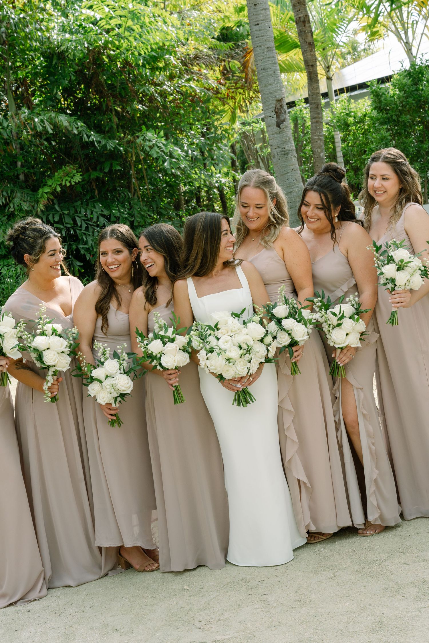 Bride in white gown poses with bridesmaids in taupe dresses holding white floral bouquets outdoors.