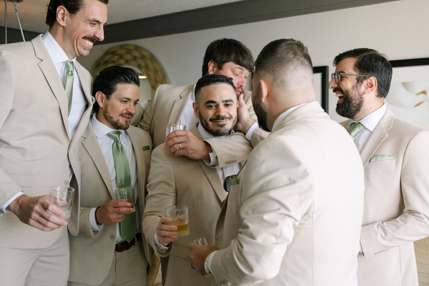 Groomsmen in light colored suits sharing drinks and laughs during wedding celebration.