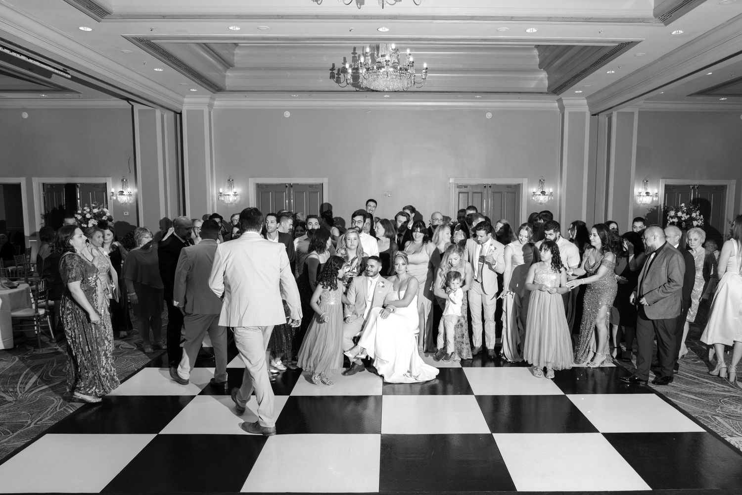 Black and white photo of people dancing on a checkered ballroom floor at an elegant event with a chandelier overhead.