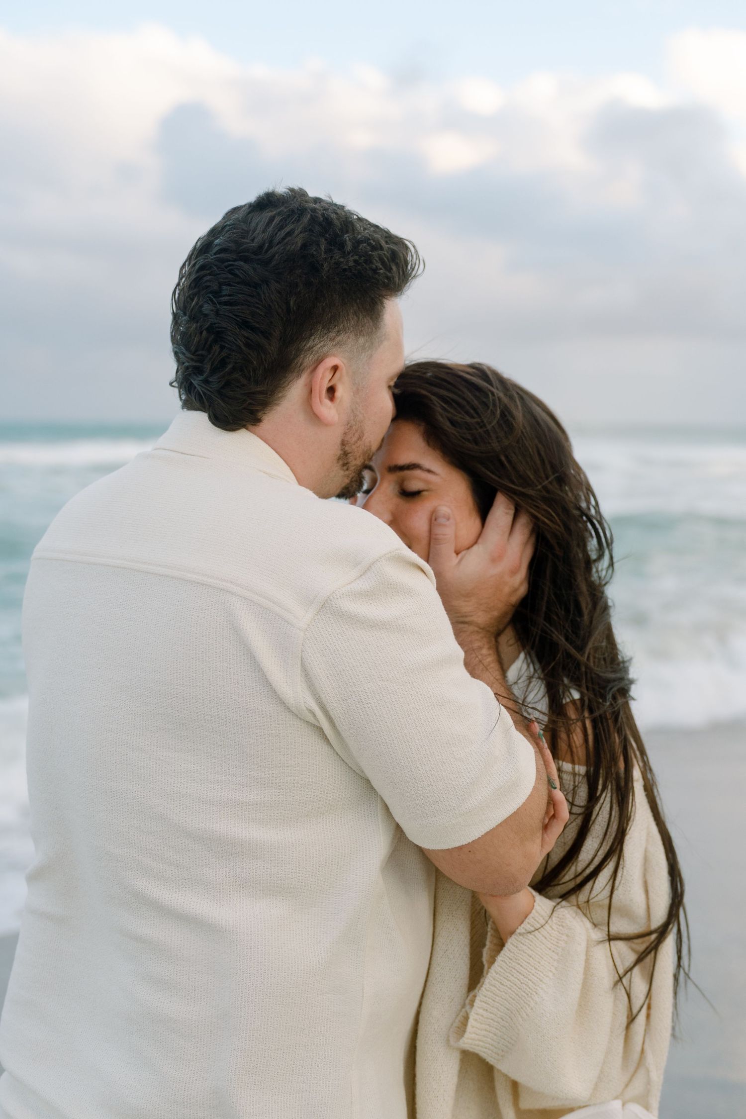A romantic couple embraces and kisses on a cloudy beach with ocean waves in the background.