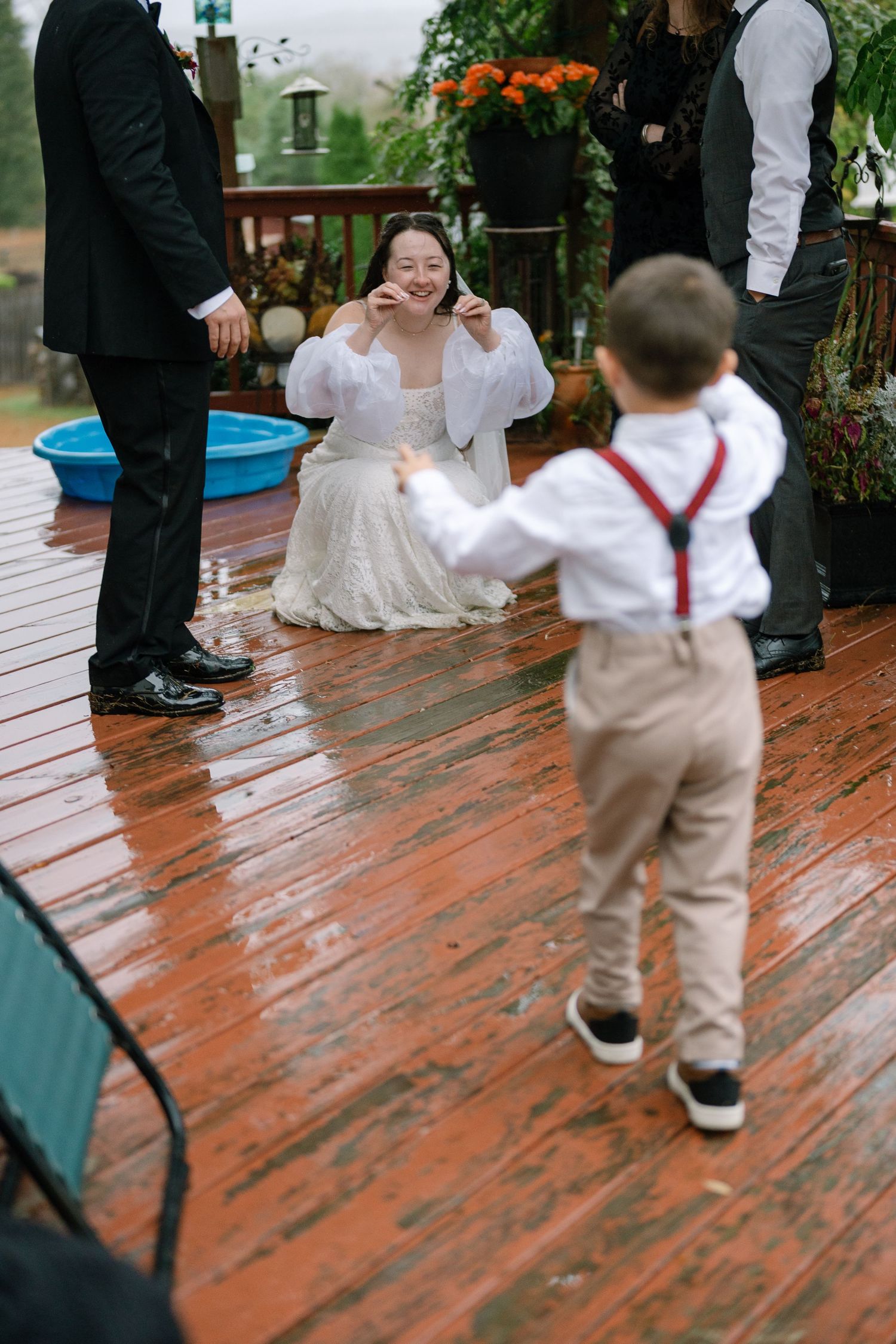 A toddler in suspenders and khakis walks across a wooden deck during a wedding celebration.