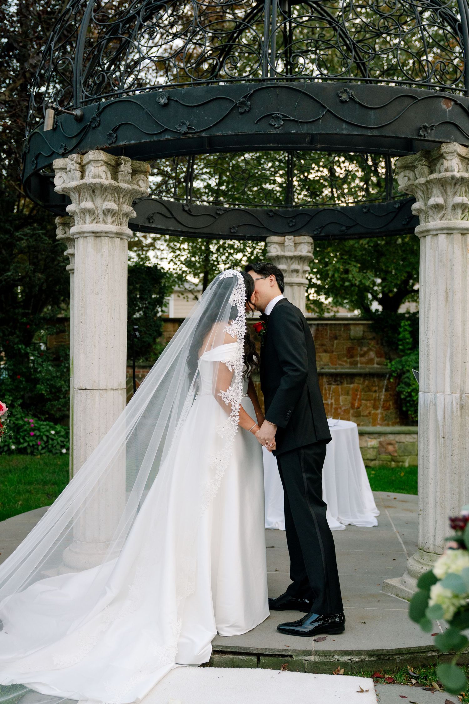 A romantic wedding ceremony takes place under an elegant white gazebo with flowing veils and pillars in an outdoor garden setting.
