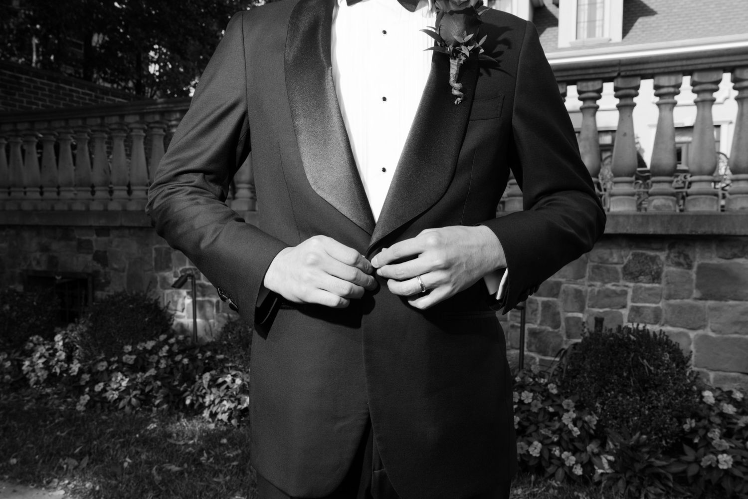 Black and white photo of someone in a formal tuxedo adjusting their jacket button outdoors by stone architecture.