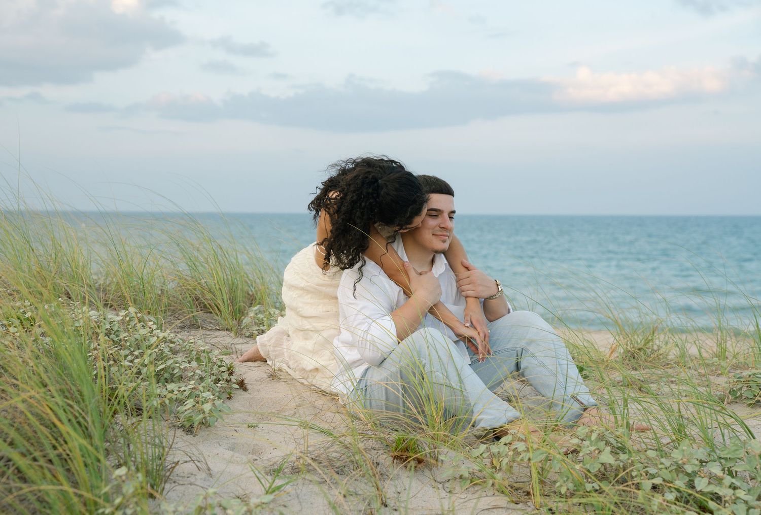 A couple sits together on a sandy beach dune with tall grass and ocean views at sunset.