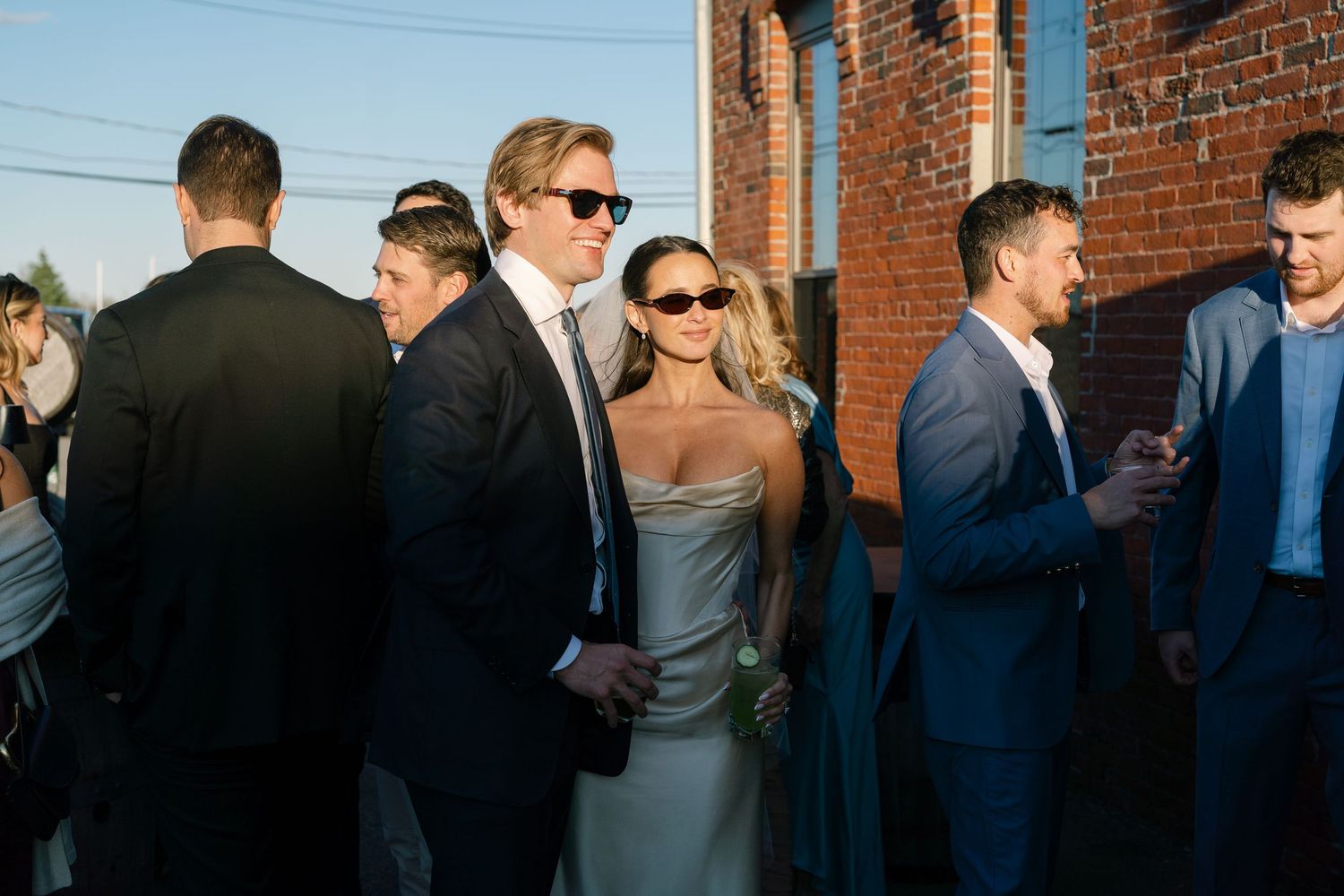 Elegantly dressed wedding guests in suits and a silver gown mingle outdoors near a brick building at a stylish event.
