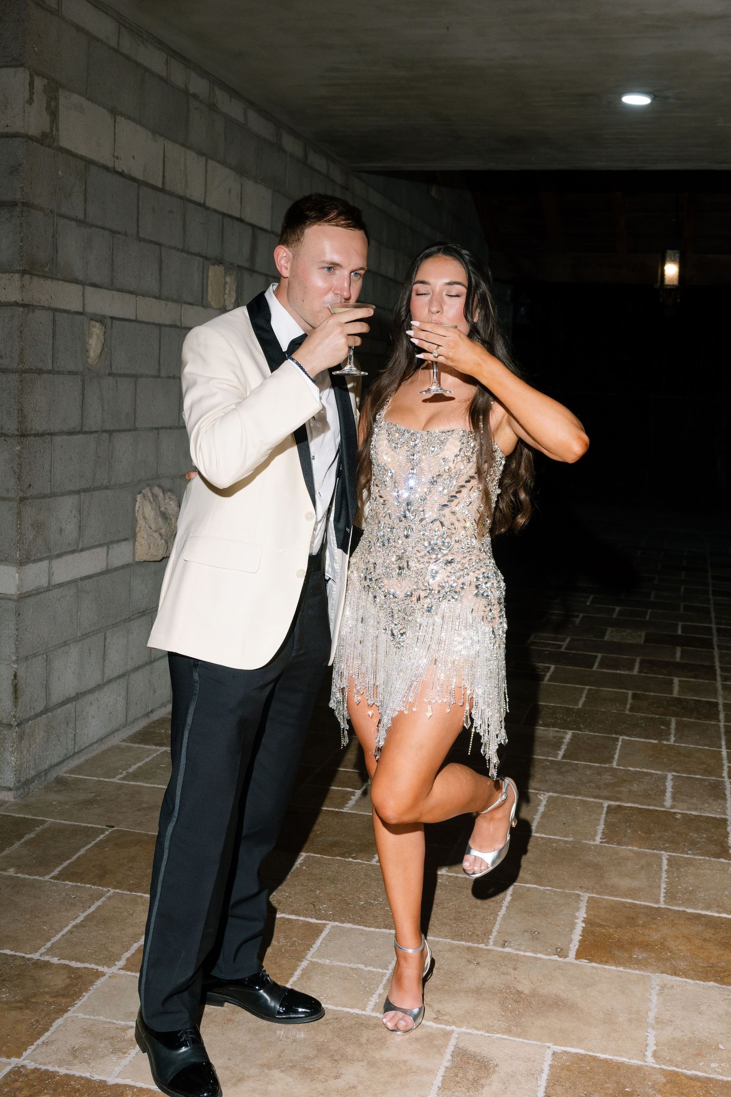A couple in formal attire drinks champagne together in a dimly lit stone corridor at a wedding reception.
