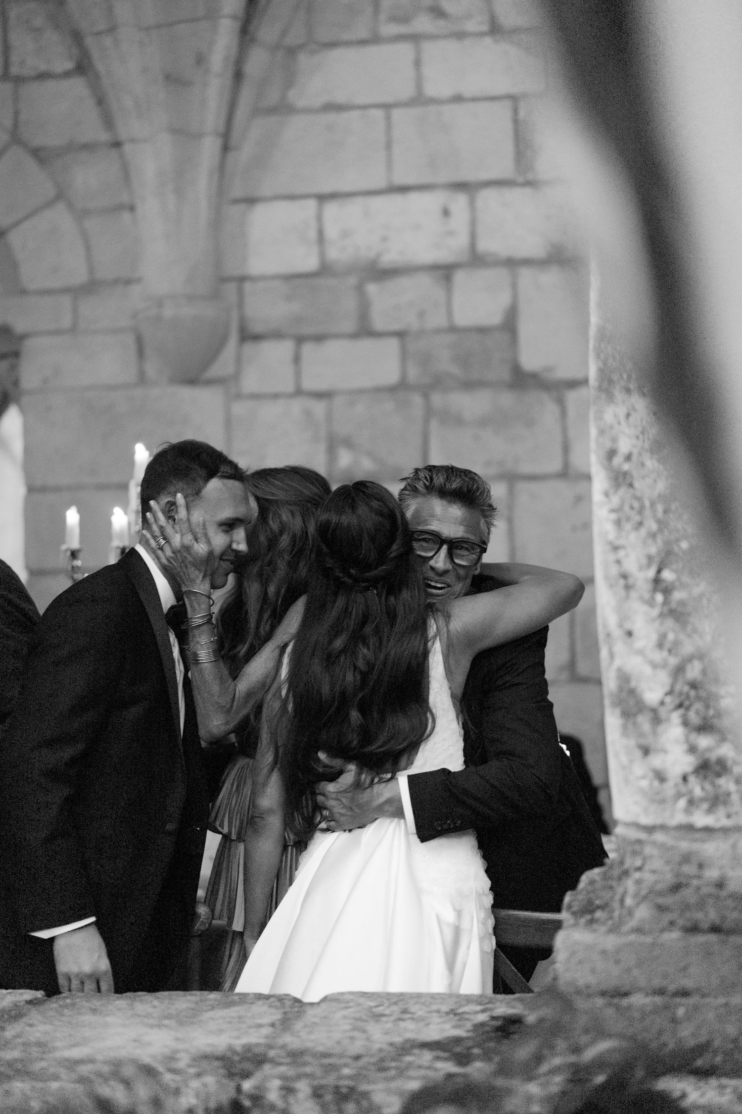 Bride hugs smiling guest in glasses near candlelit stone wall in black and white wedding photo.