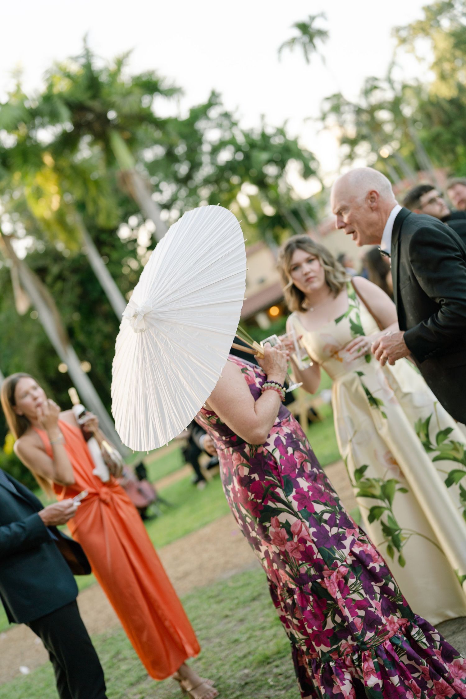 Wedding guests mingle outdoors, woman in floral dress holds white parasol among tropical palms.