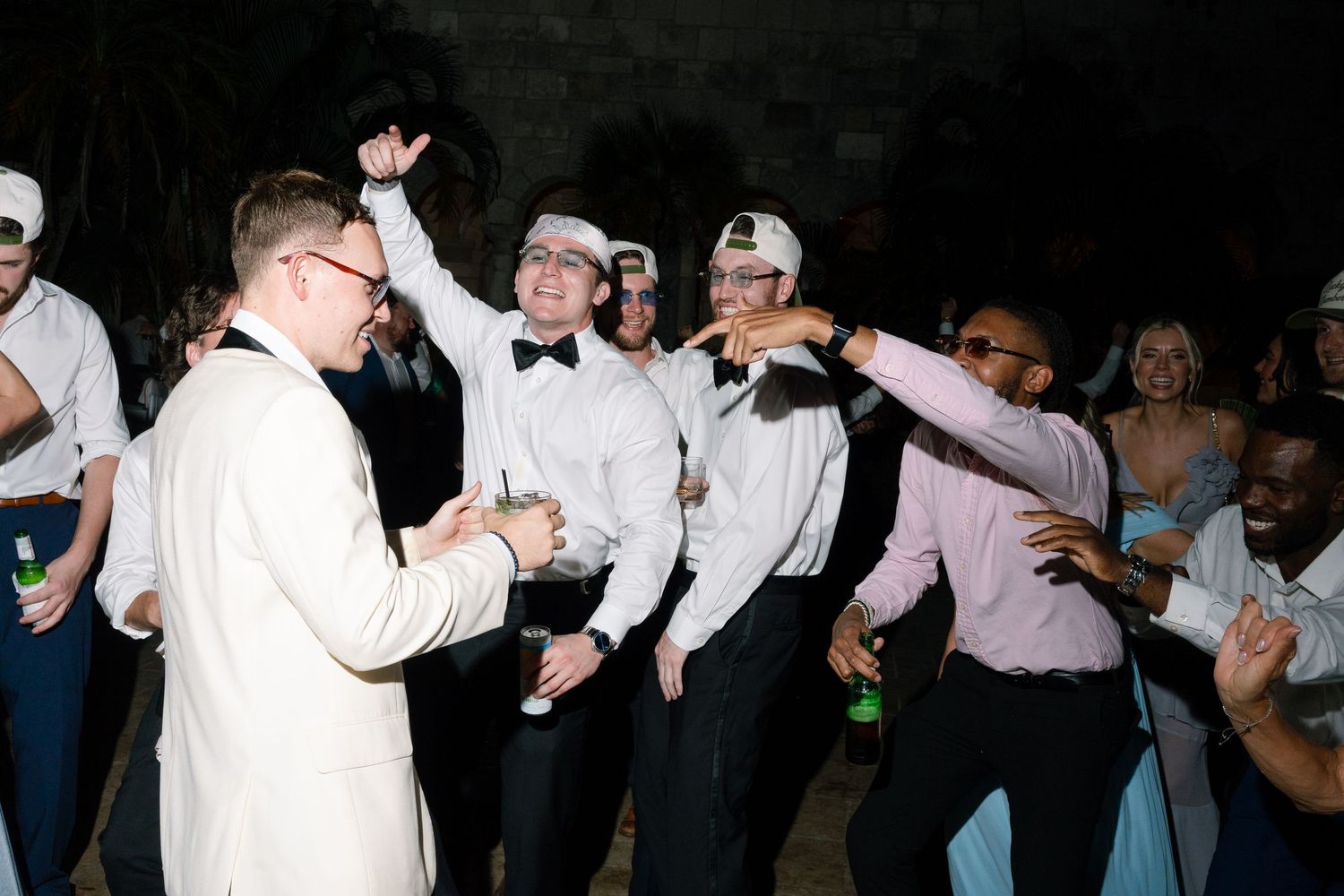 Groom in white suit dancing with groomsmen in caps at lively outdoor wedding reception party.
