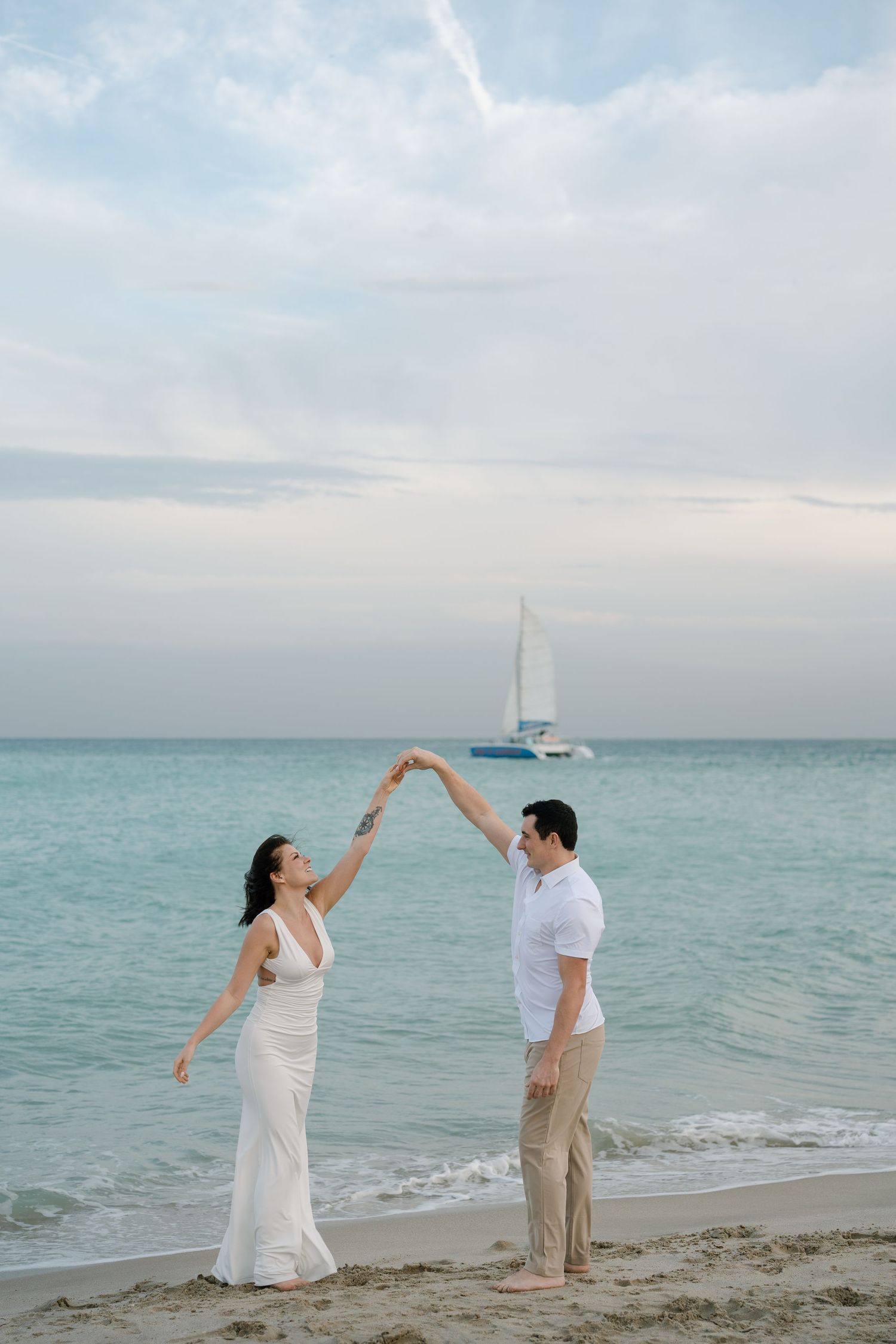 A couple dances playfully together on a sandy beach with calm ocean waters and a sailboat in the distance.