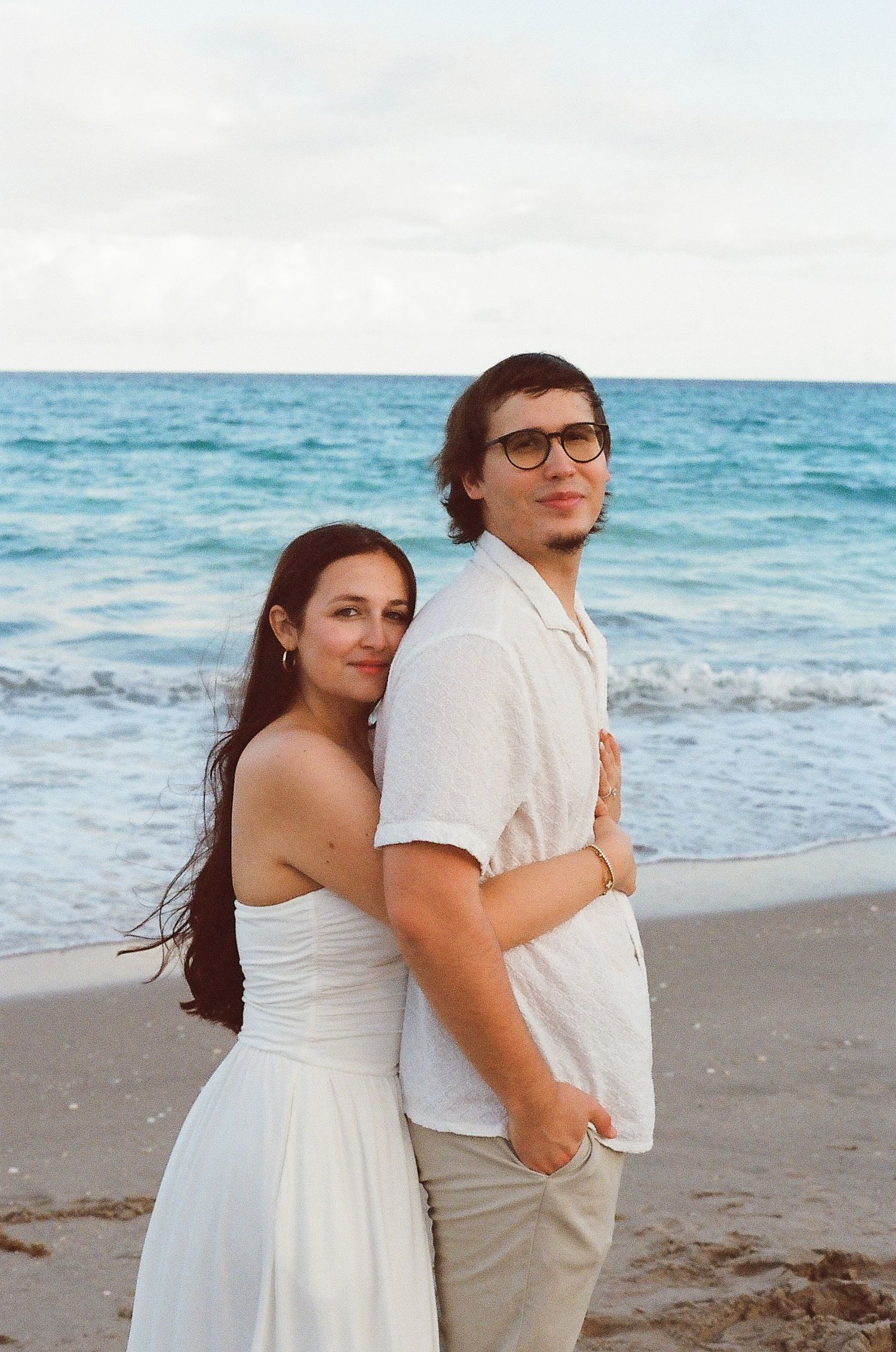A couple poses together on a scenic beach with waves and cloudy skies in the background.