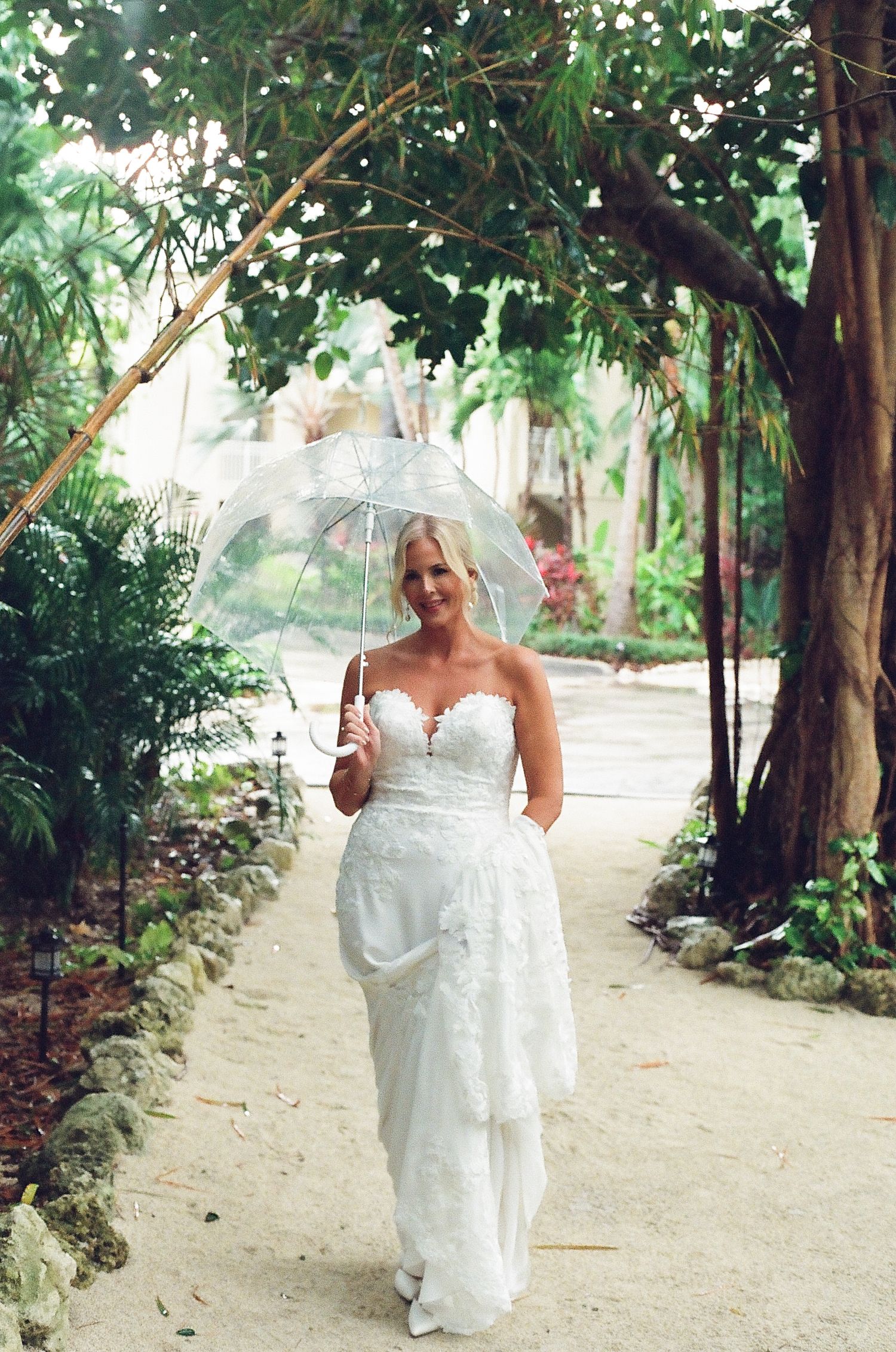A bride in a white lace wedding dress walks along a garden path holding a clear umbrella on her special day.