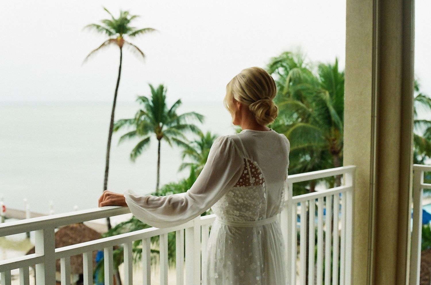 A person in white stands on a balcony overlooking tropical palm trees and ocean views.