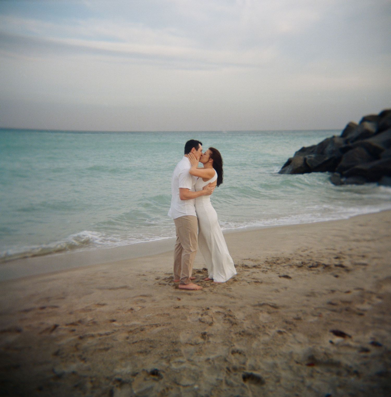 Couple shares tender moments on sandy beach with ocean waves and cloudy sky in background.