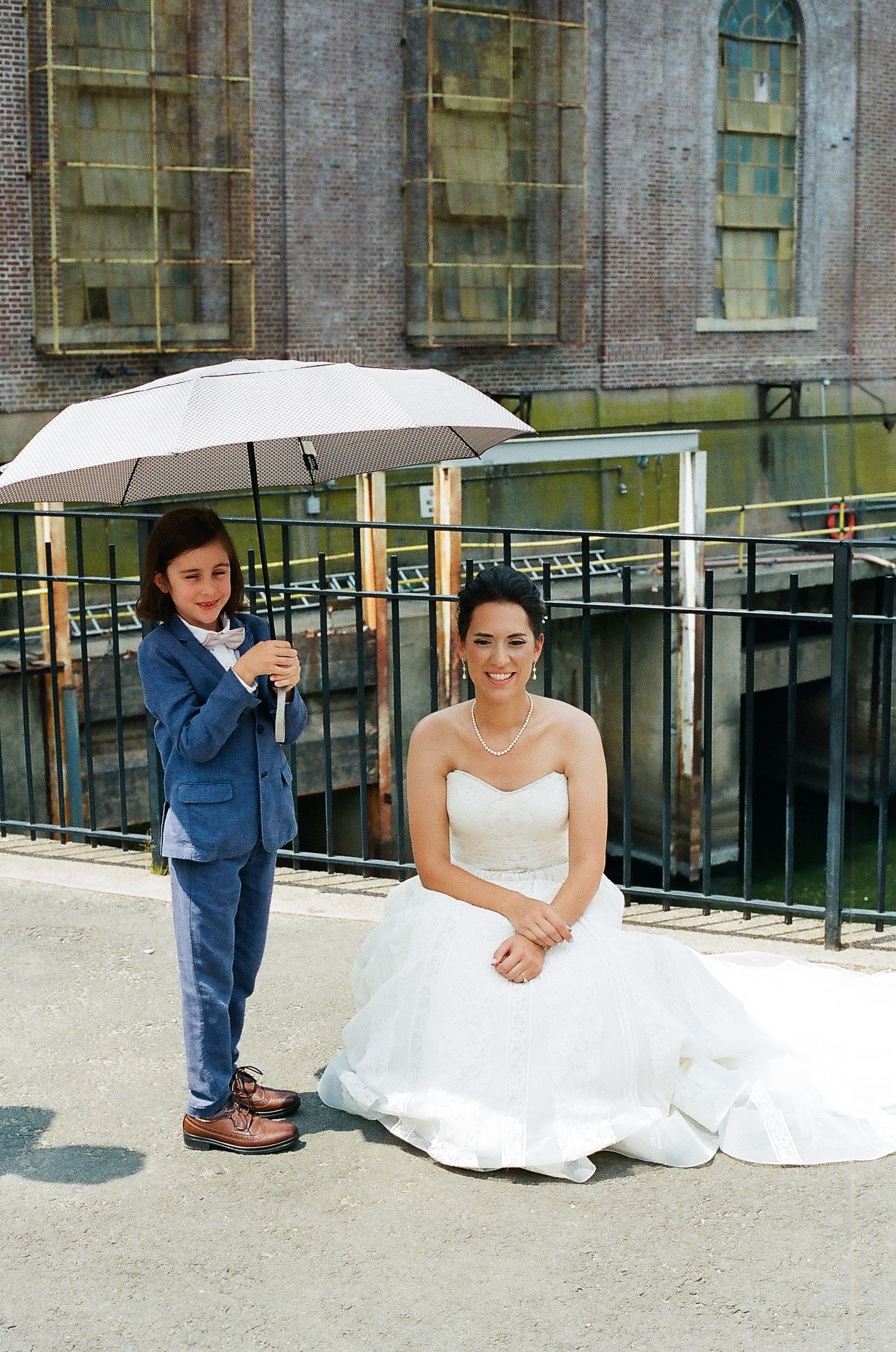 A playful wedding photoshoot on a balcony with a white parasol and elegant white dress against a brick building.