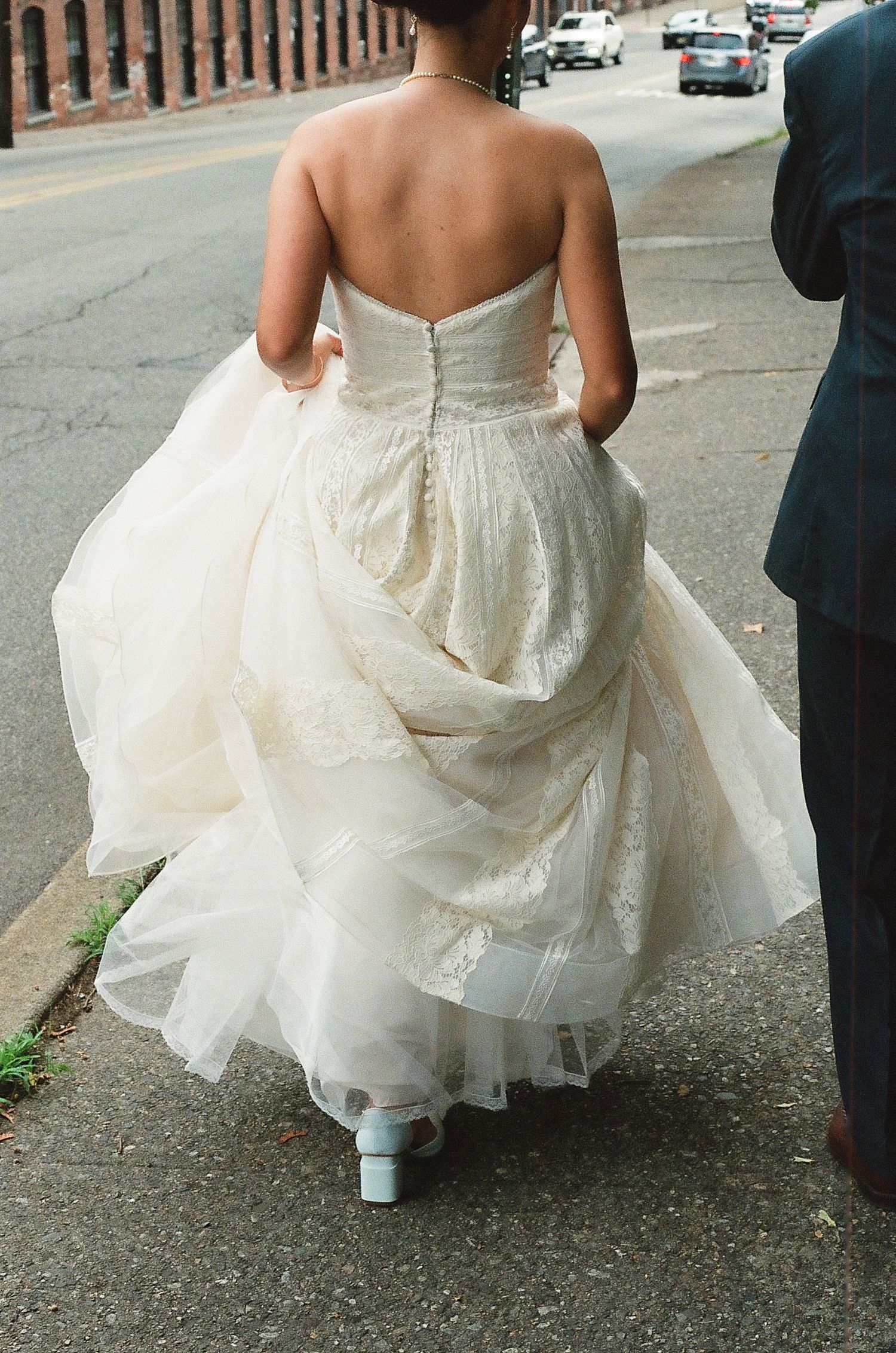 Back view of flowing white wedding dress train and heels on a city street.