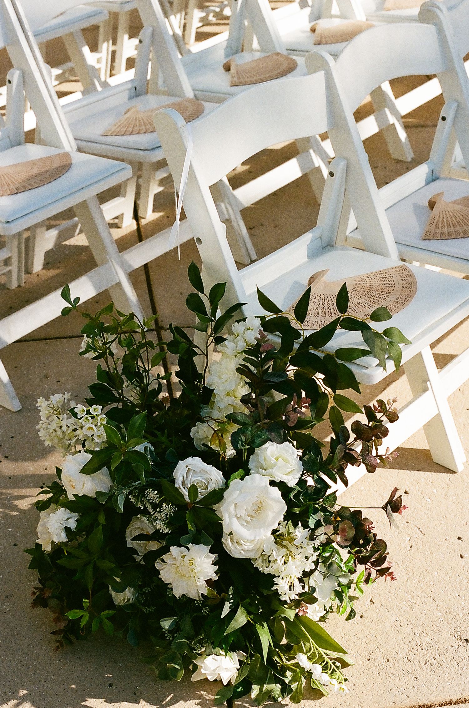 White folding chairs decorated with lush floral arrangements line up at an outdoor wedding ceremony.