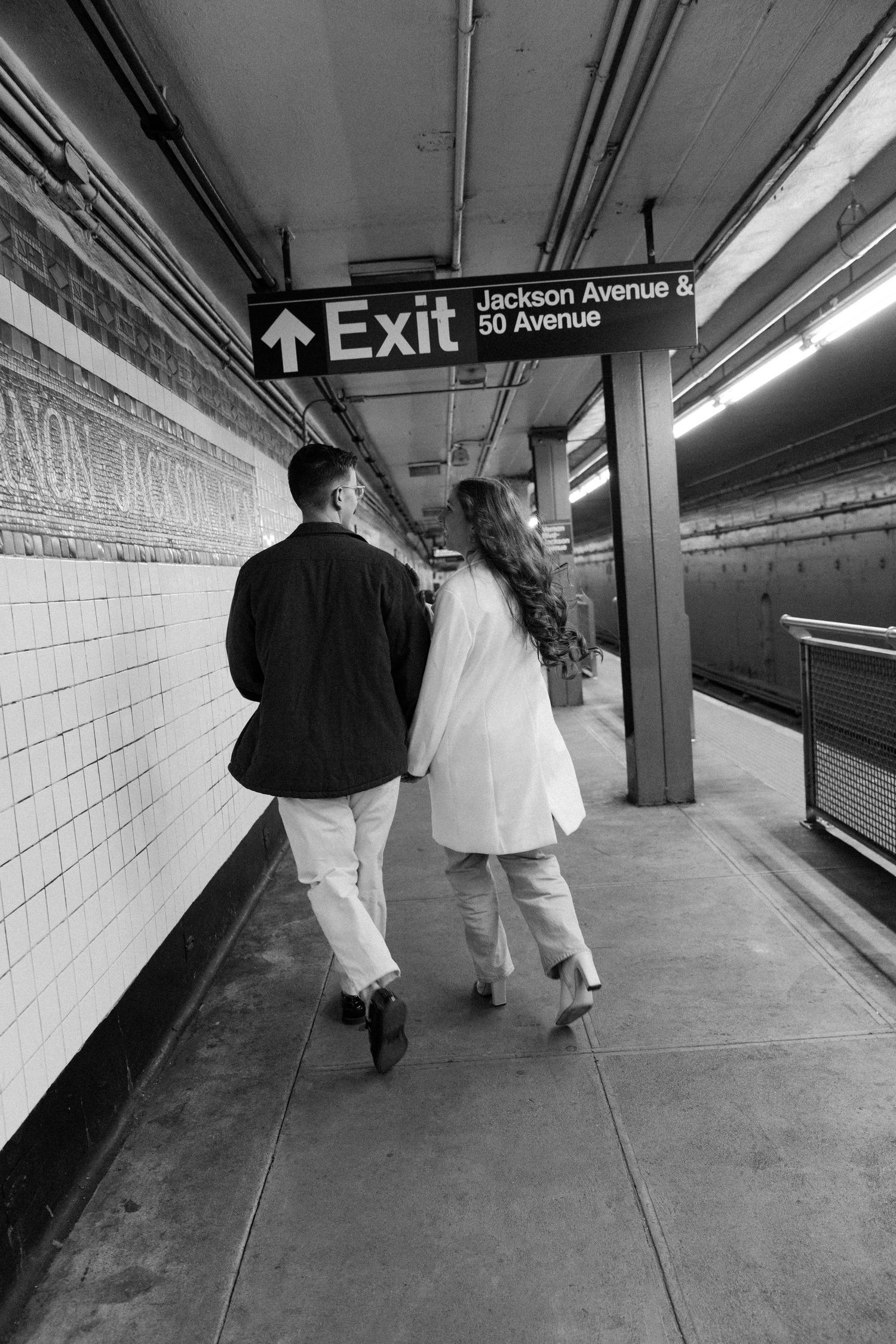 People walking along an empty subway platform at Jackson Avenue station in black and white.