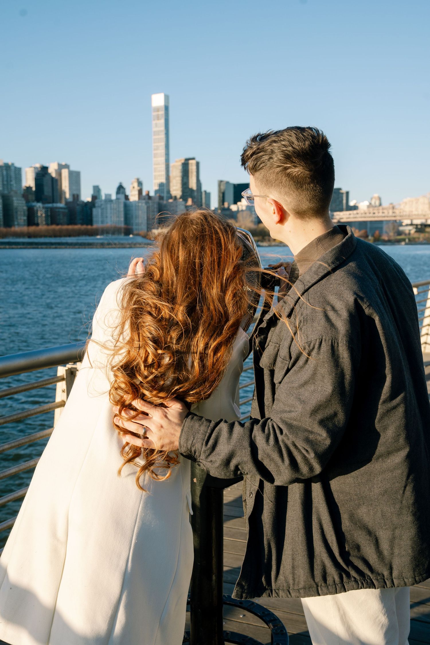 Two people in contrasting black and white outfits lean against railing overlooking city skyline.