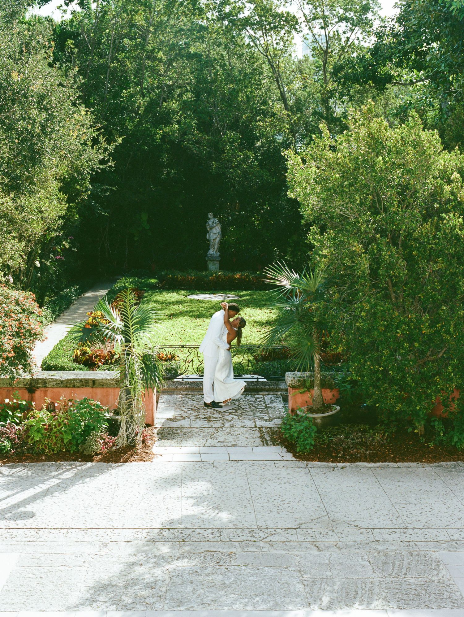 A stone paved garden pathway leads through lush greenery with decorative brick pillars and potted plants.