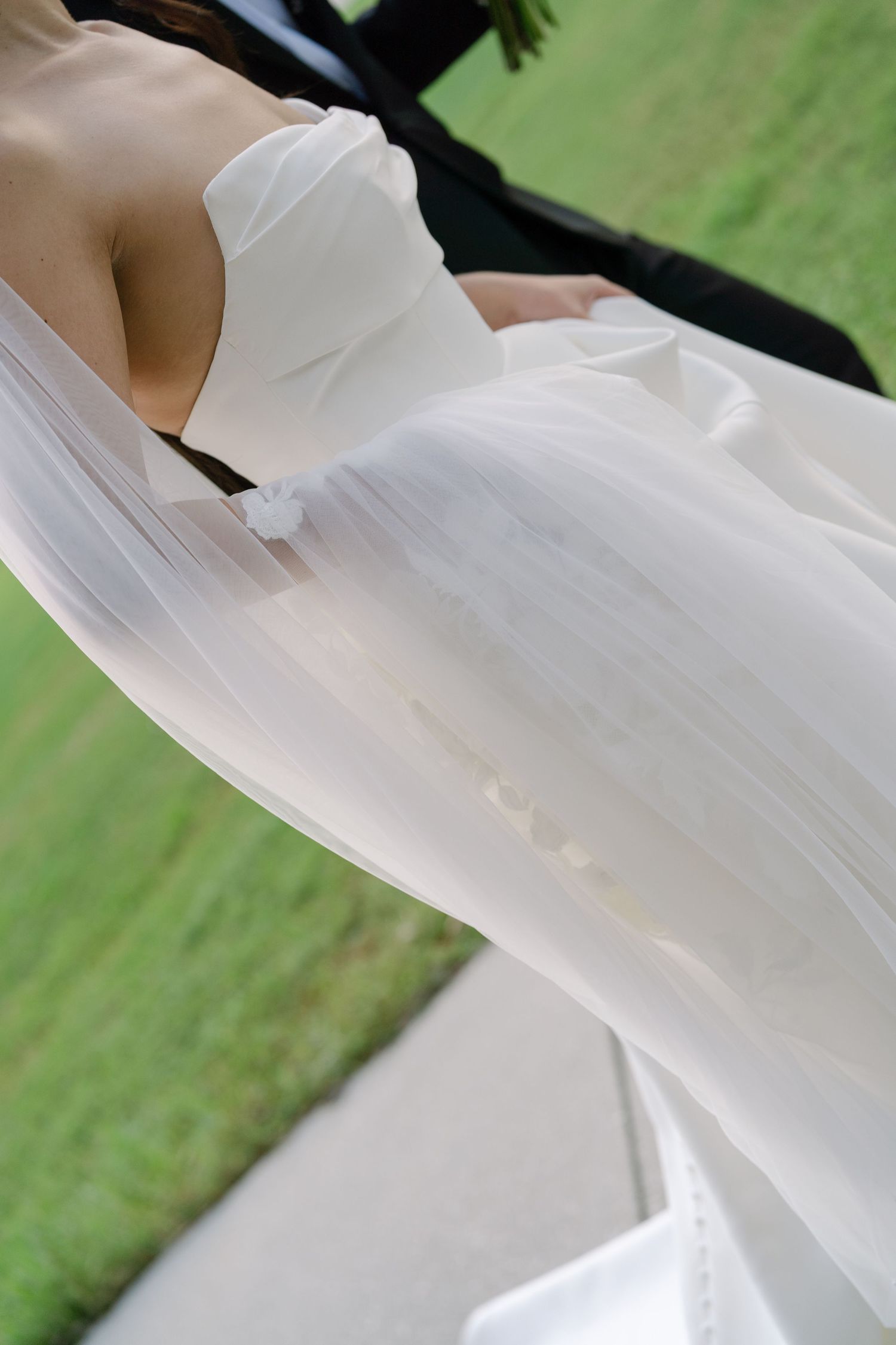 Close-up of a bride in a flowing white strapless wedding gown with sheer veil, standing outdoors on green grass.