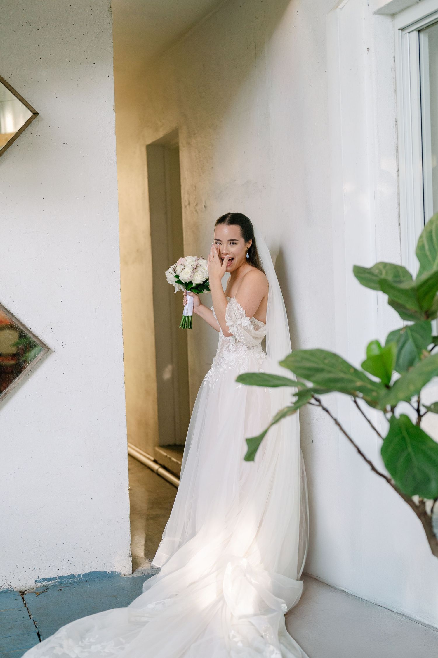 Bride in white lace gown holds bouquet, hand over mouth in surprise, leaning against wall by window.