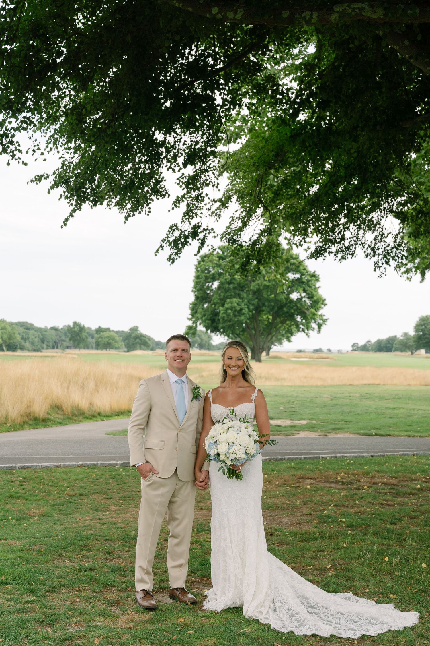 Bride and groom pose together under a large tree on a golf course, she holds a white bouquet in her lace wedding gown.