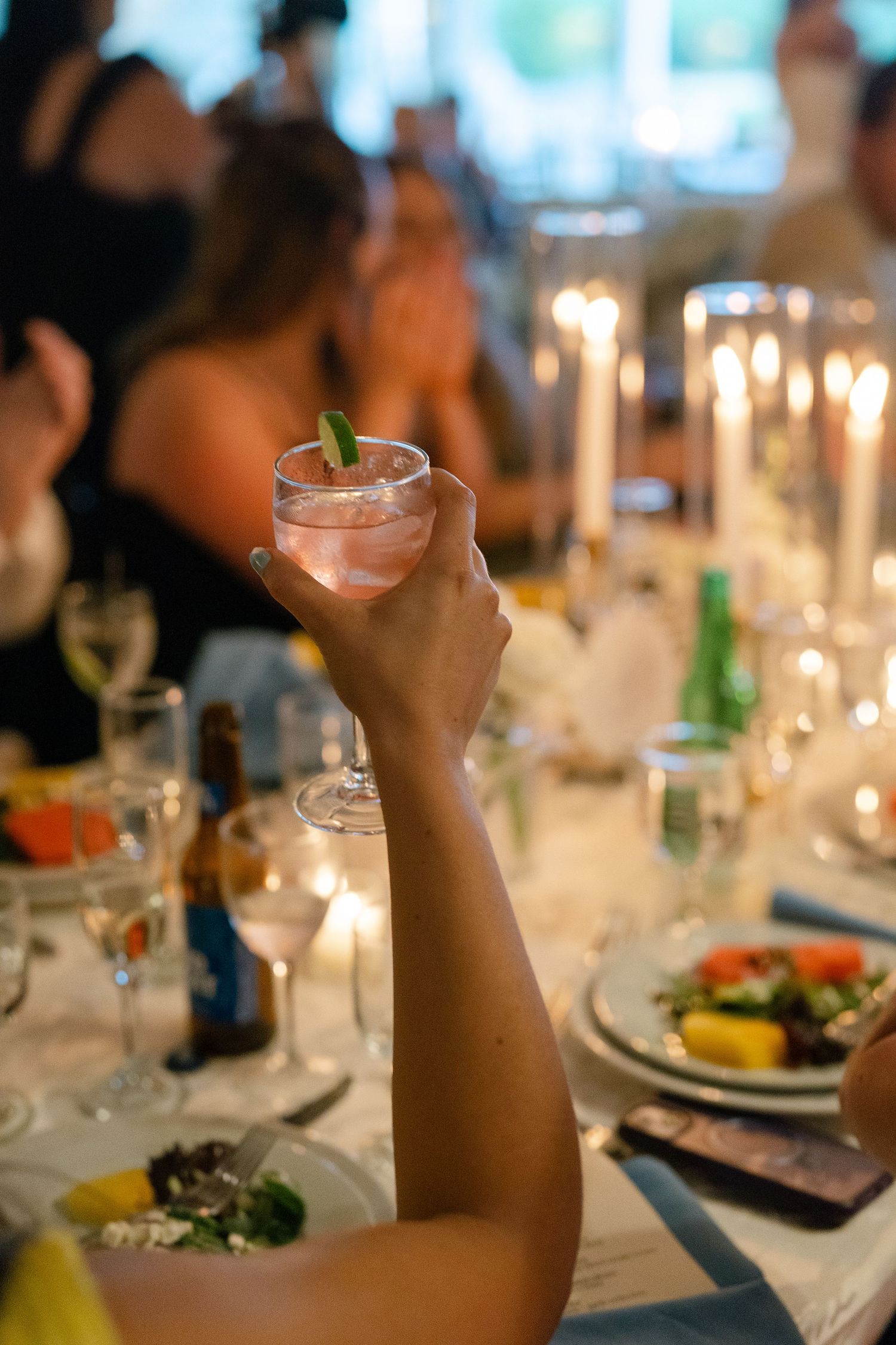 Wedding guest raises a pink cocktail in a toast at a candlelit reception dinner table.