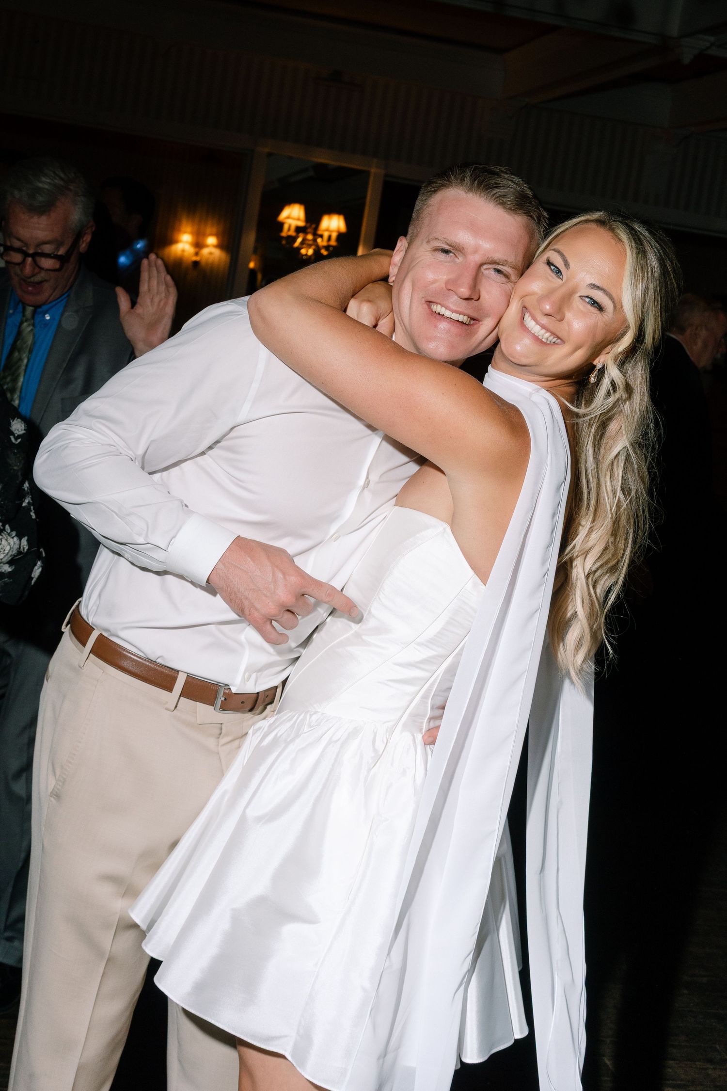 Smiling couple in white outfits embracing at a wedding reception on a dance floor.