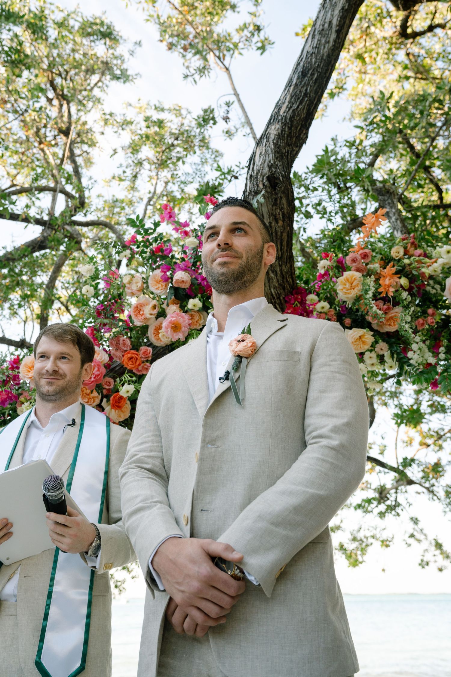 A wedding ceremony under a floral arch with pink and peach blooms against a backdrop of trees.