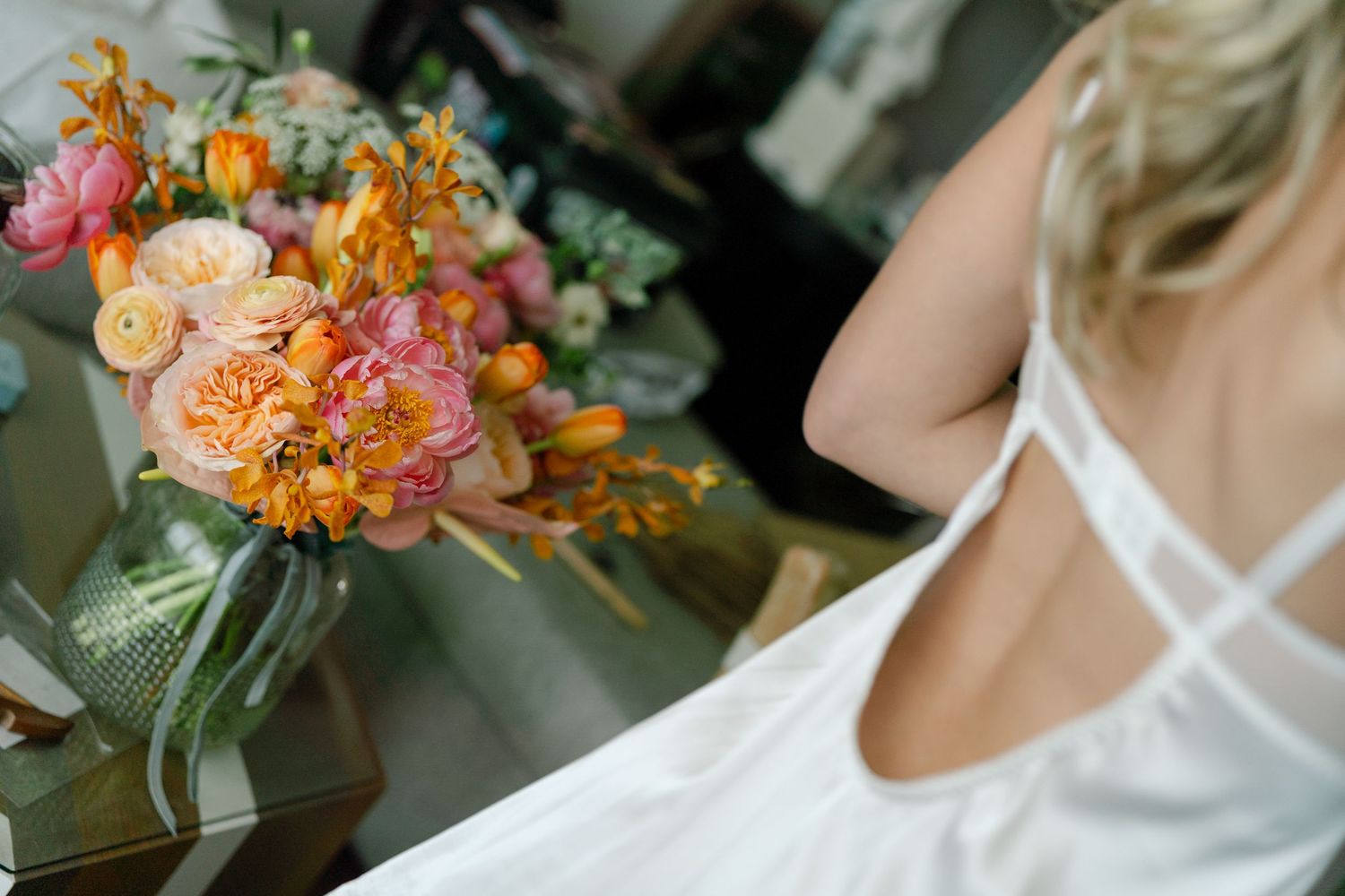 A vibrant bouquet of peach, orange and pink flowers sits on a table next to someone wearing a white strappy dress.