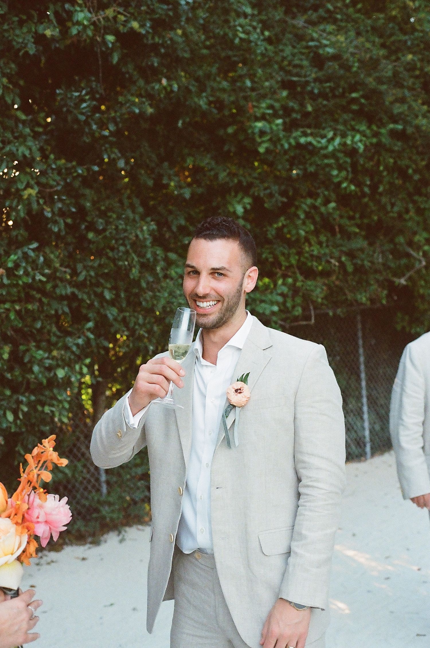 Groomsmen in light gray suits share a candid moment outdoors against a green hedge backdrop.