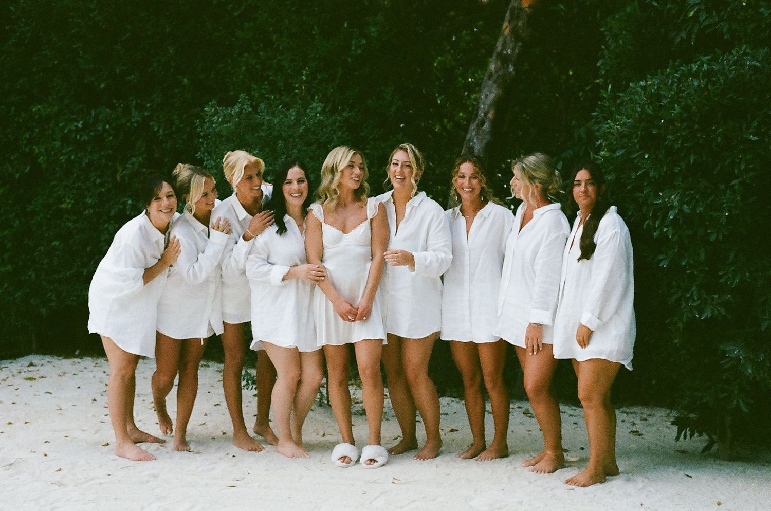Row of people in white clothing sitting together against a dark green hedge background.