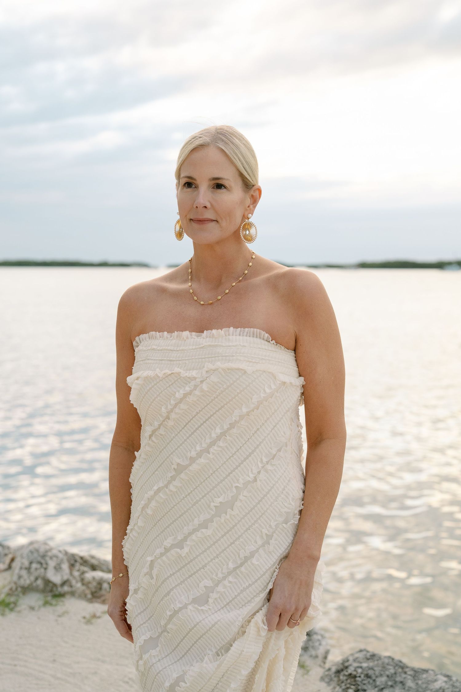 Beachside portrait session in white strapless dress with diagonal details against shimmering water at sunset.