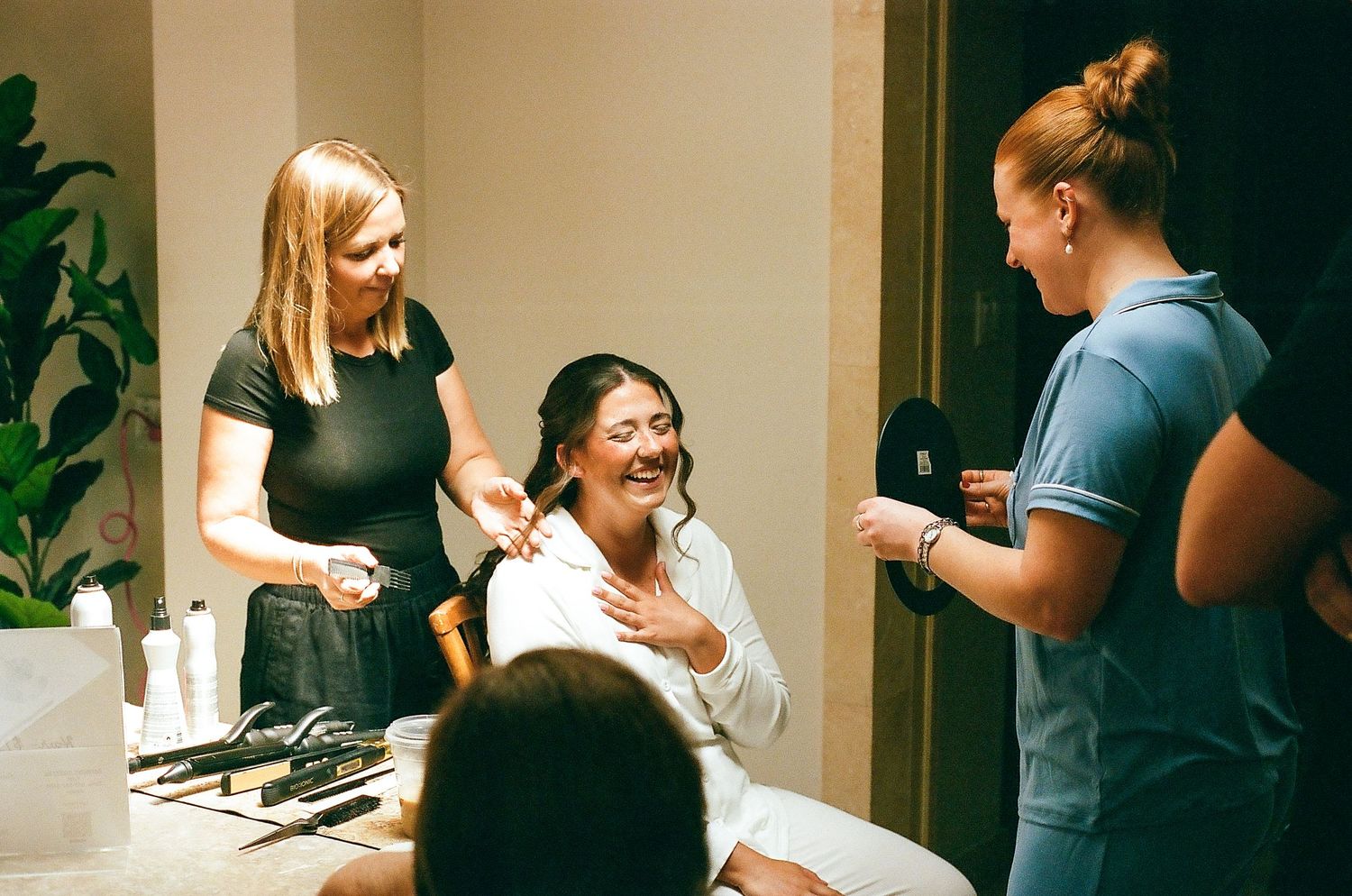 Bride smiling in white robe getting hair and makeup done by two stylists in a bright salon.