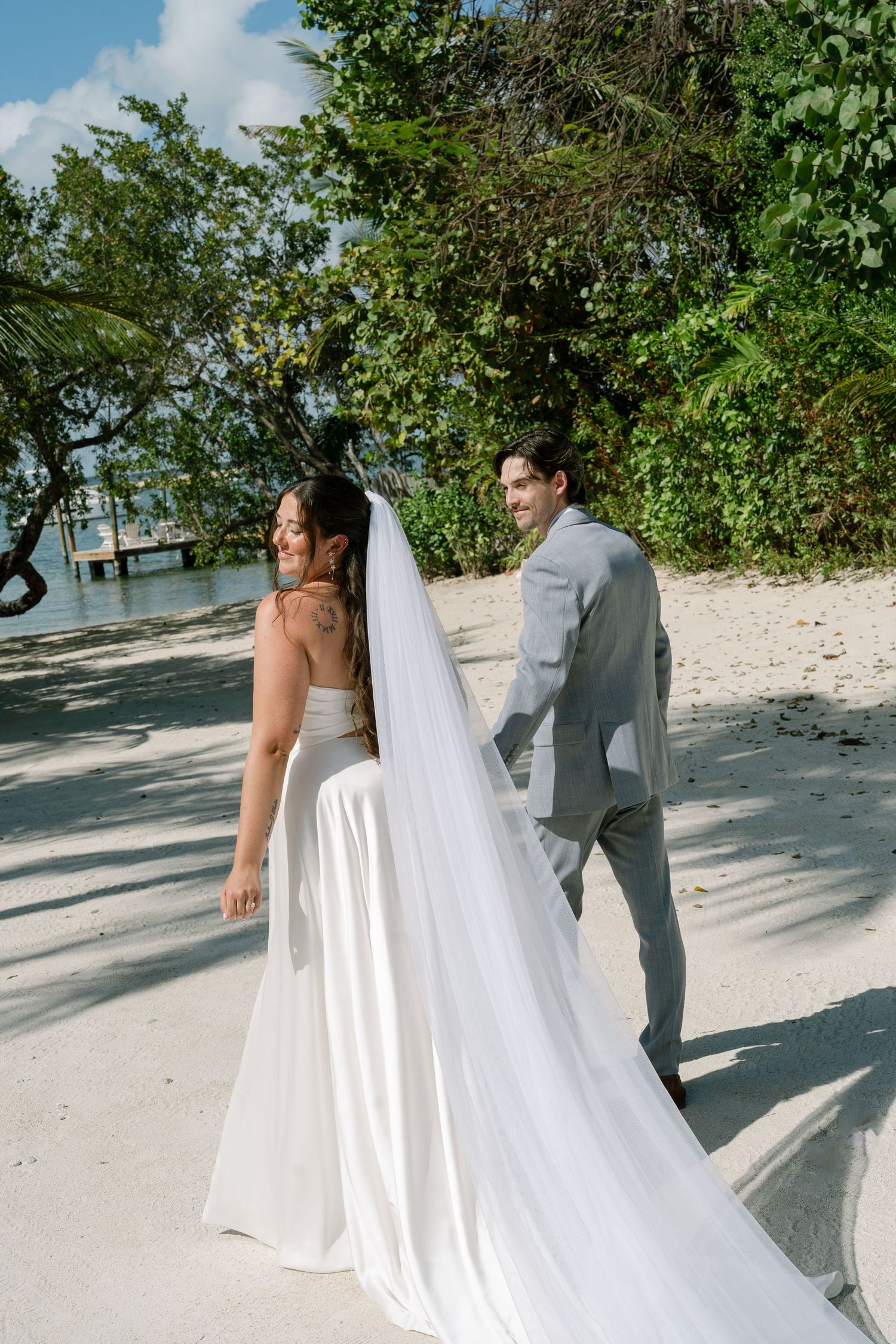 A bride and groom stand together on a beach, surrounded by lush greenery.