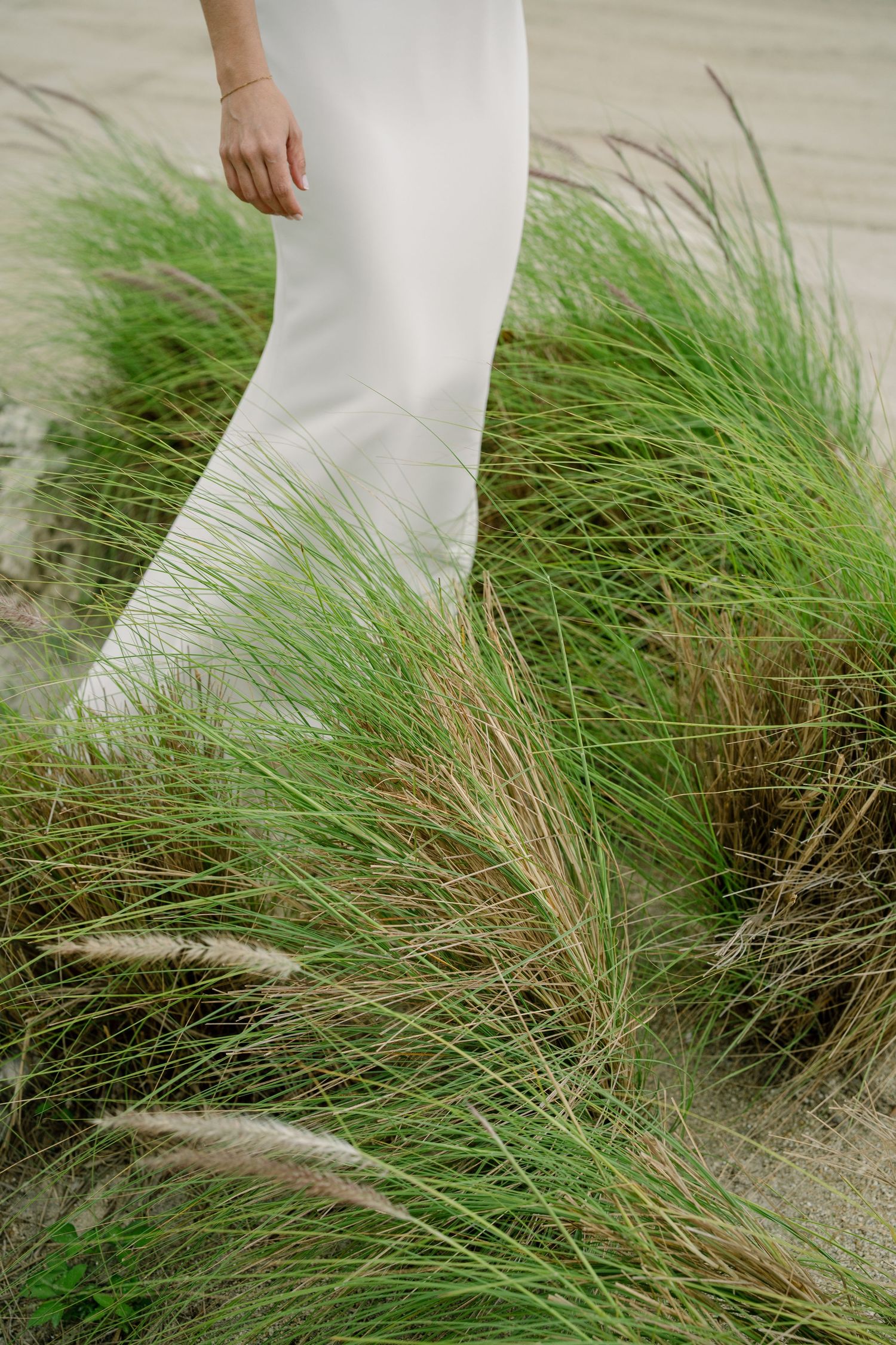 Person wearing white pants walking through tall beach grass swaying in the coastal breeze.