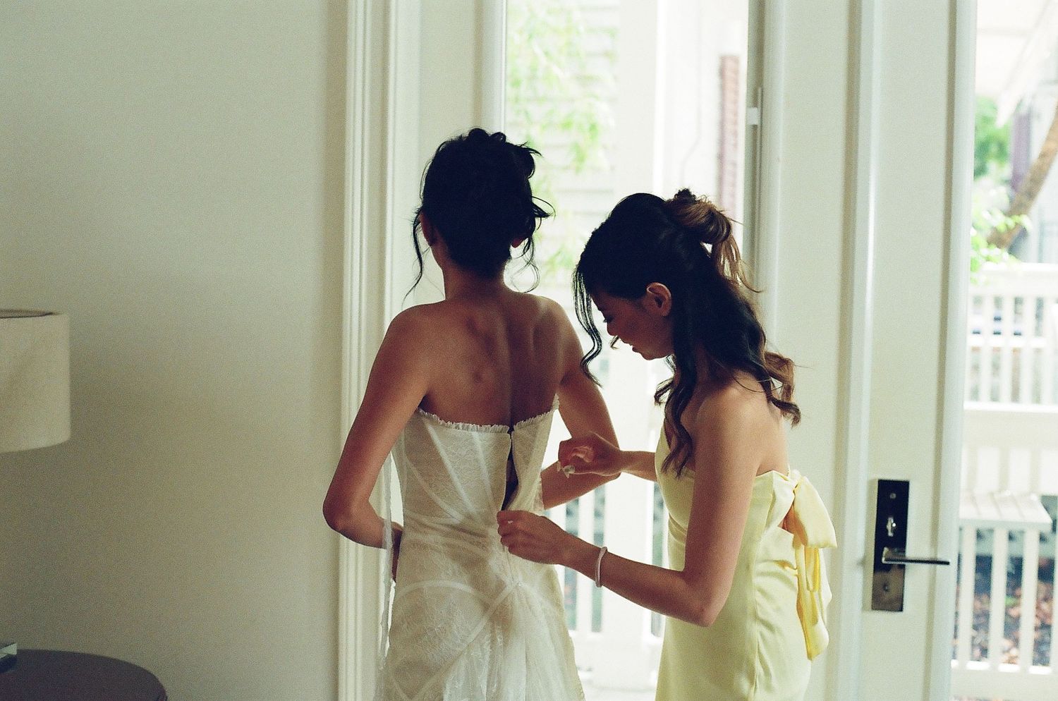 Bridesmaid in yellow dress helping bride button up white wedding gown near glass door.