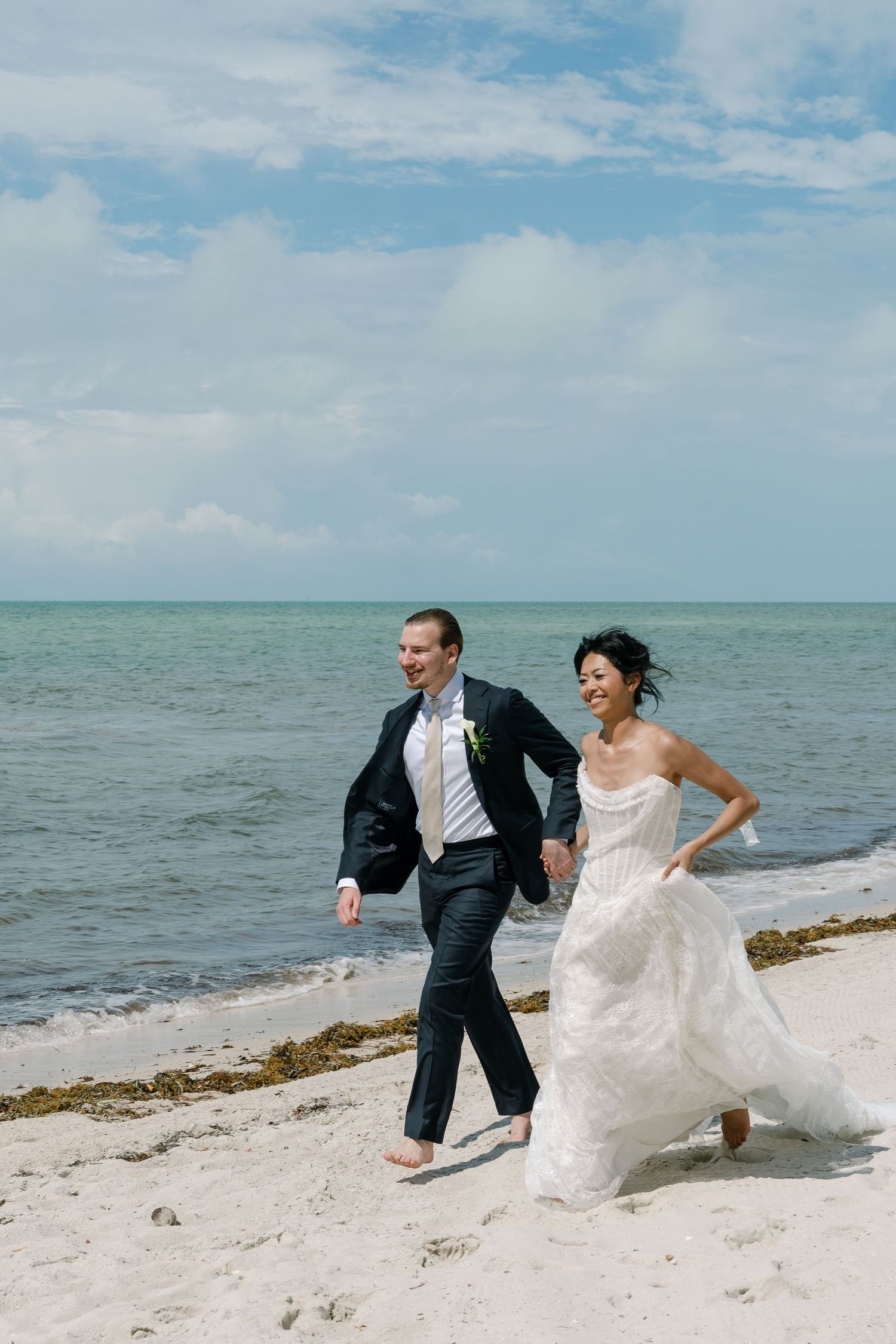Bride and groom running barefoot on a sandy beach in wedding attire with turquoise ocean waves in the background.