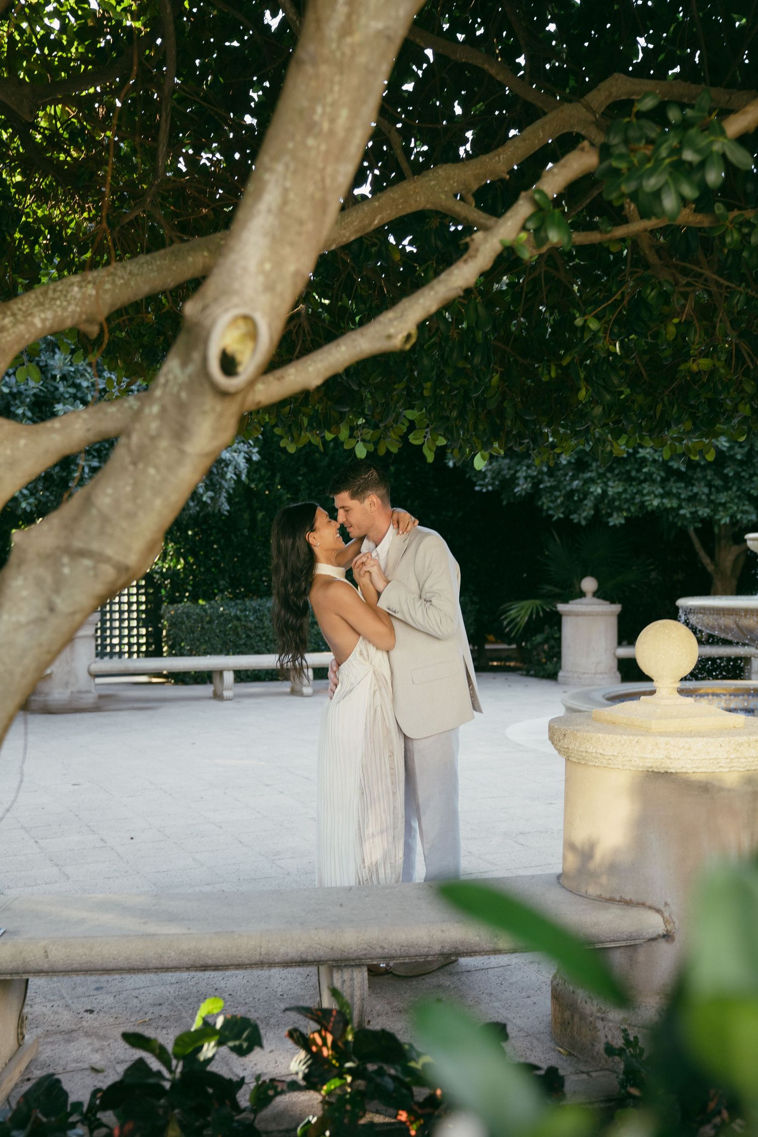 A couple shares intimate romantic moments on a terrace with tree branches overhead and stone architectural elements.