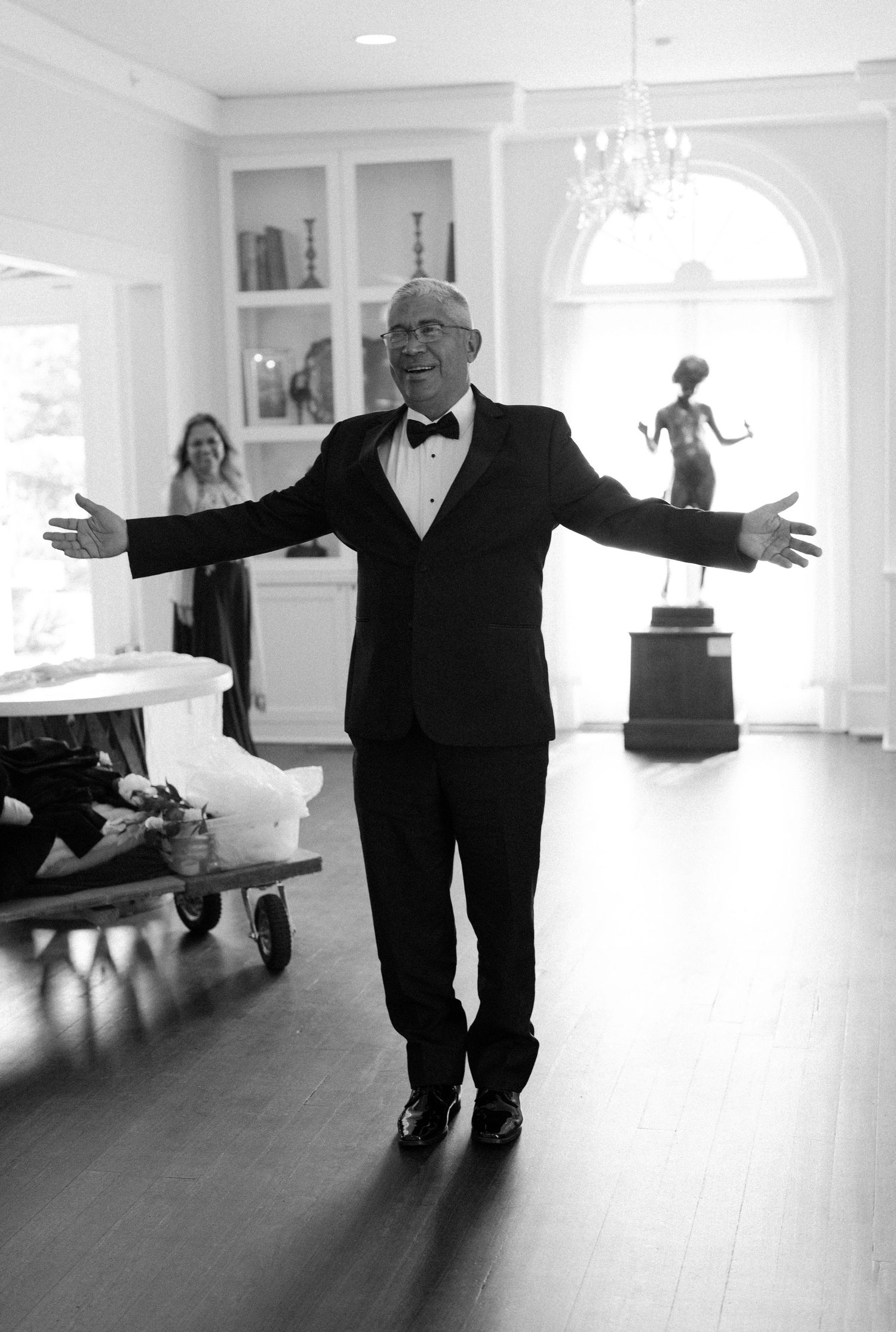 Black and white photo of an elderly person in a tuxedo with arms outstretched in a bright room with hardwood floors.