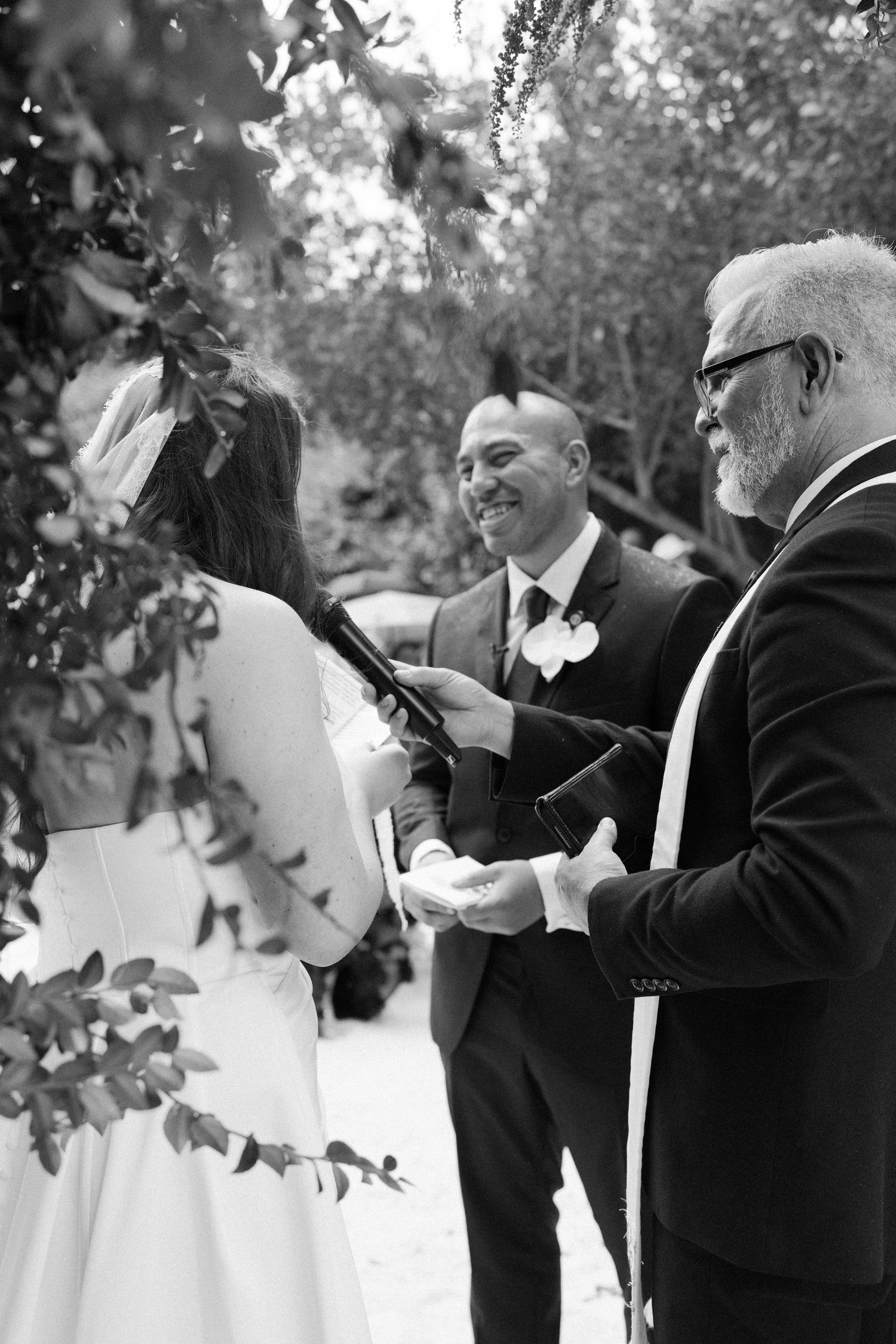 Black and white wedding ceremony photo showing an officiant reading during an outdoor garden ceremony.