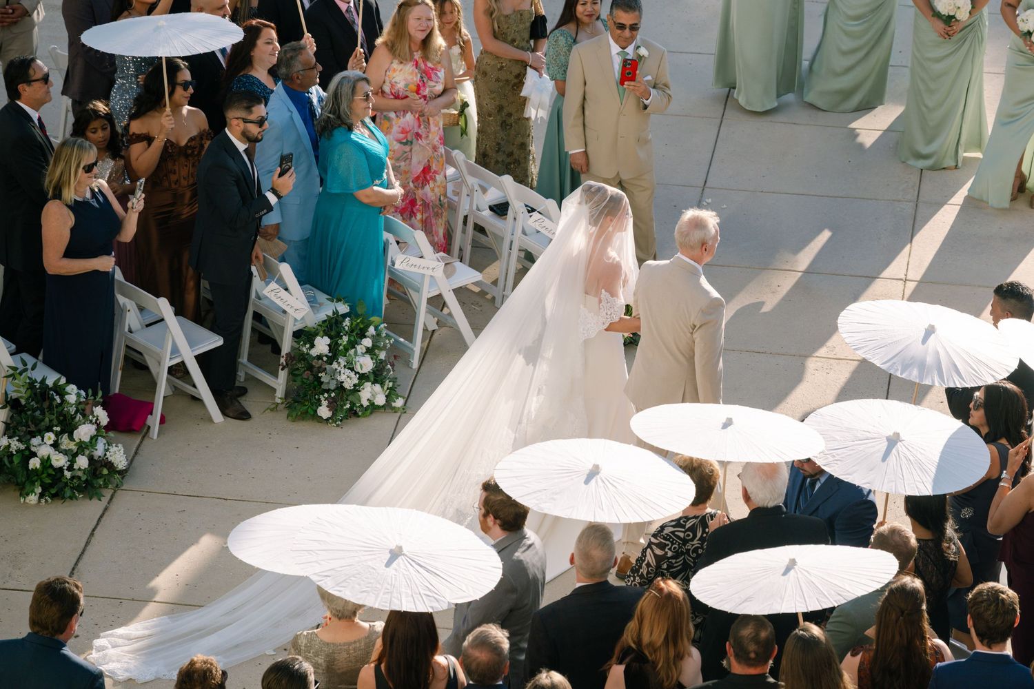 Wedding ceremony guests holding white parasols during an outdoor celebration.