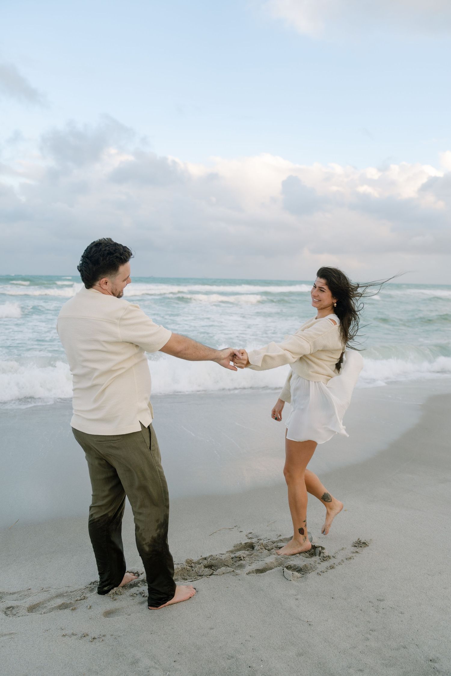 Two people playfully hold hands and dance together on a cloudy beach with waves crashing in the background.