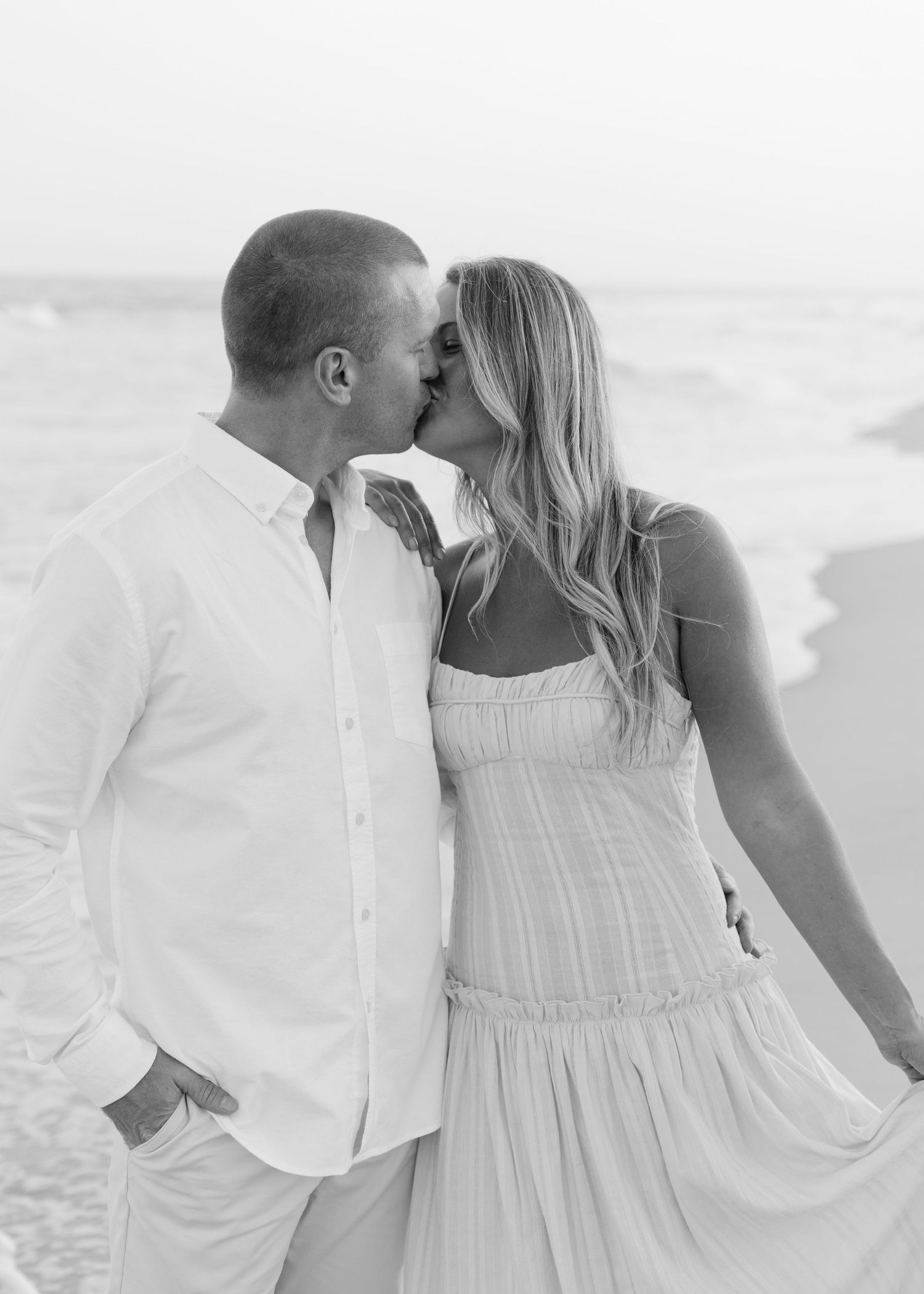 A romantic black and white photo of a couple in white outfits sharing an intimate moment on the beach.