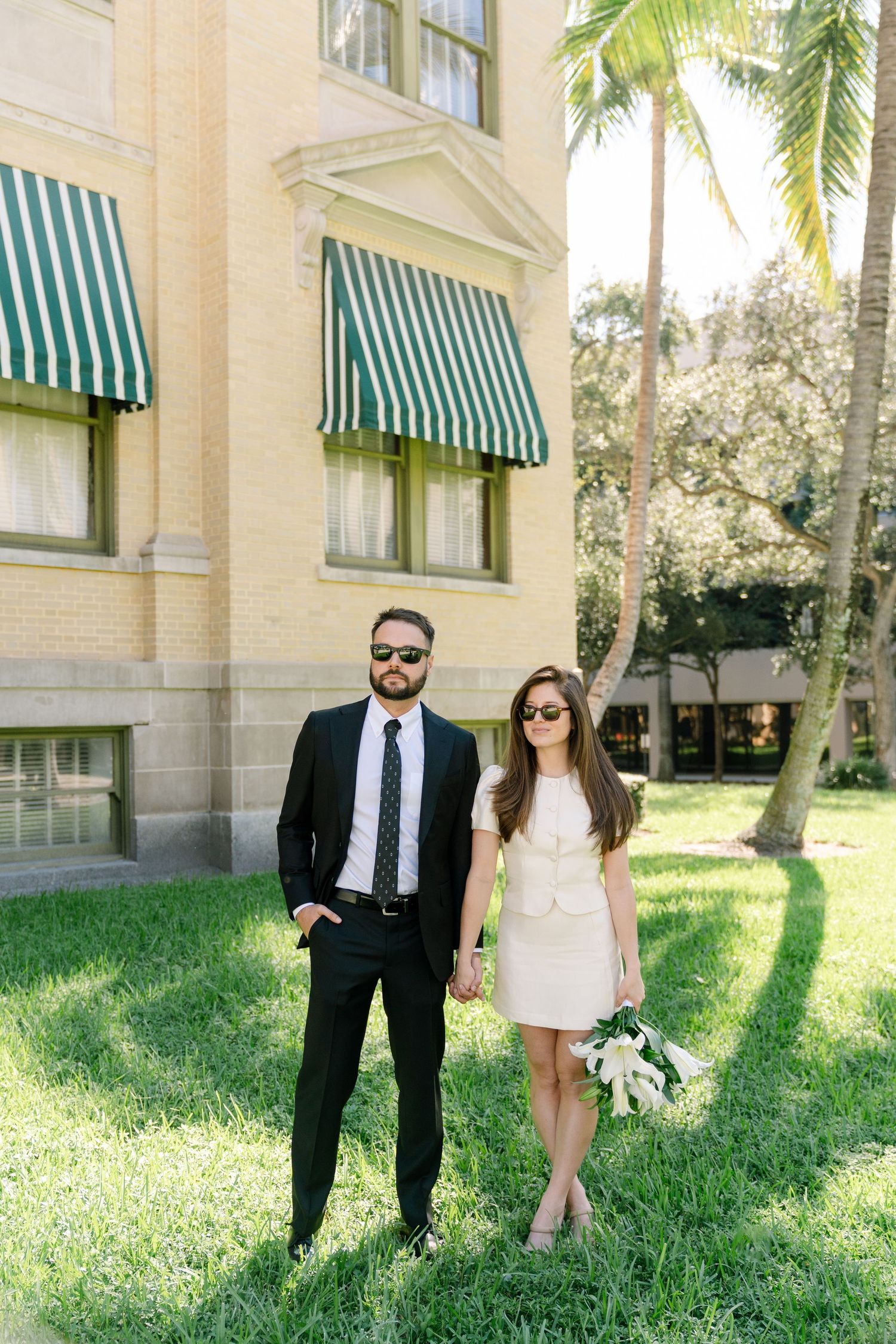A couple poses outdoors near a building with green striped awnings and palm trees on a sunny day.