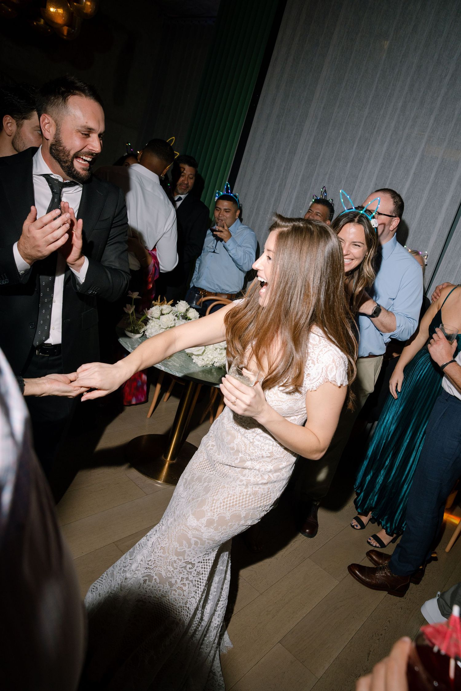 Guests dancing and celebrating at a wedding reception with green curtains and wooden flooring.